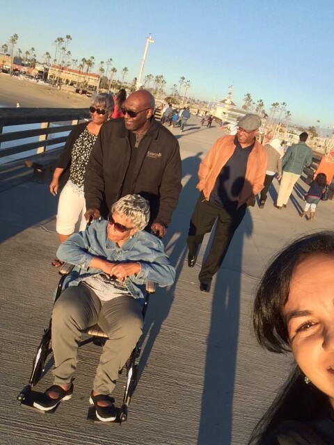Group of people walking on a sunny seaside pier; an elderly woman in a wheelchair is pushed by a man in a dark jacket while others stroll behind; a partial selfie of a woman is visible in the foreground on the right.