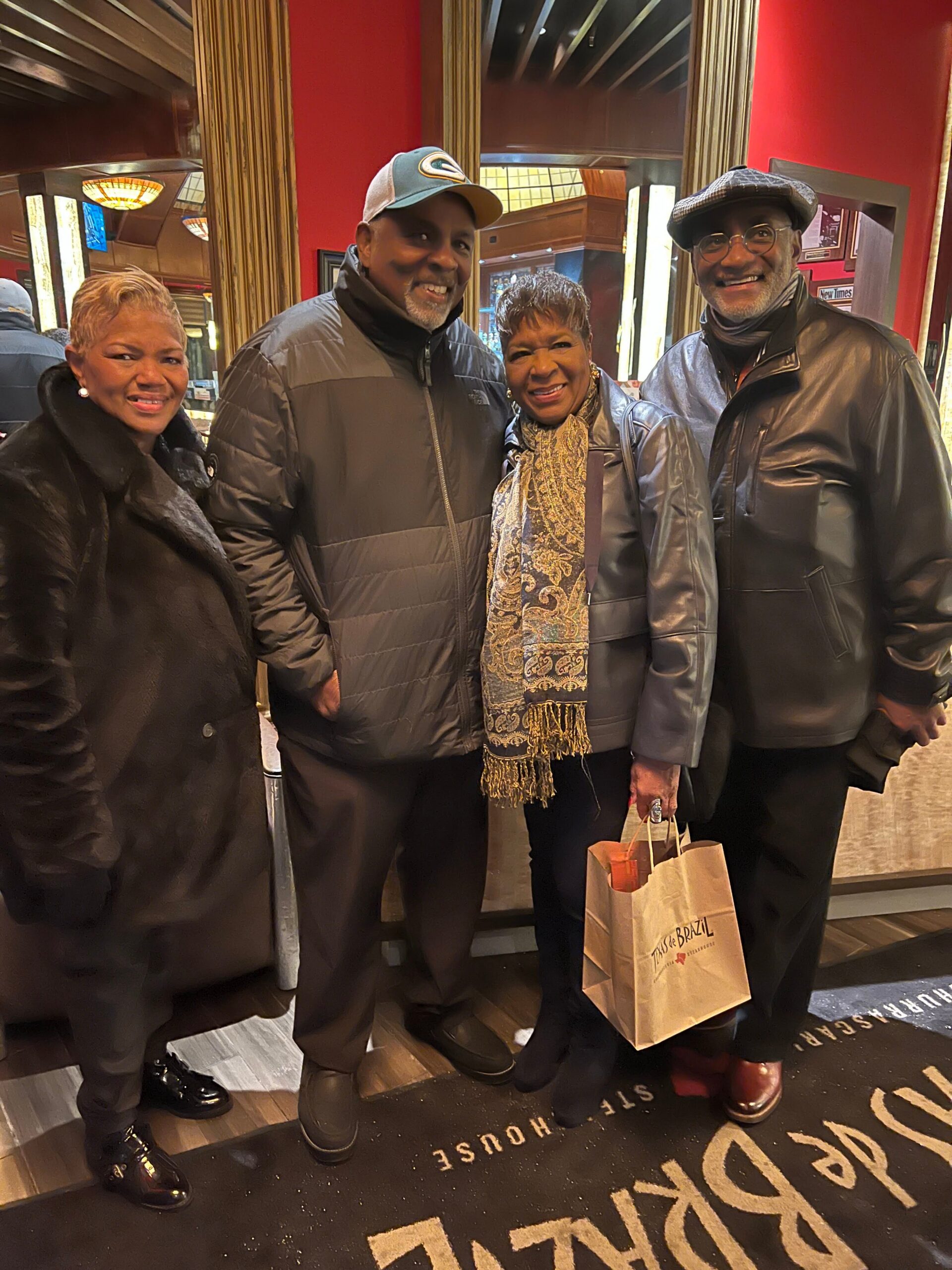 Four adults in winter jackets posing together and smiling inside a shop, one holding a brown paper bag.