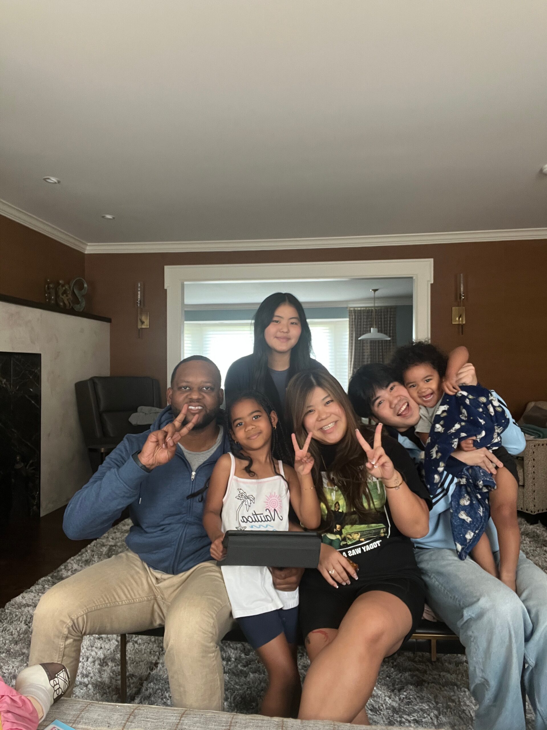 Family and friends posing on a sofa, making peace signs and smiling in a living room setting.