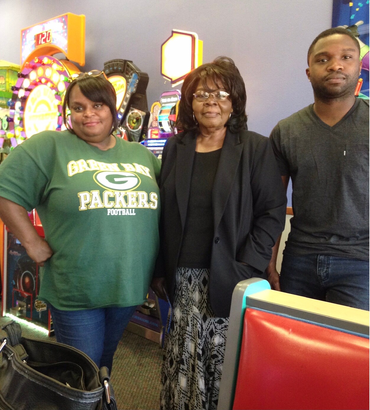 Three people pose for a photo in a game arcade, with colorful arcade machines visible in the background.