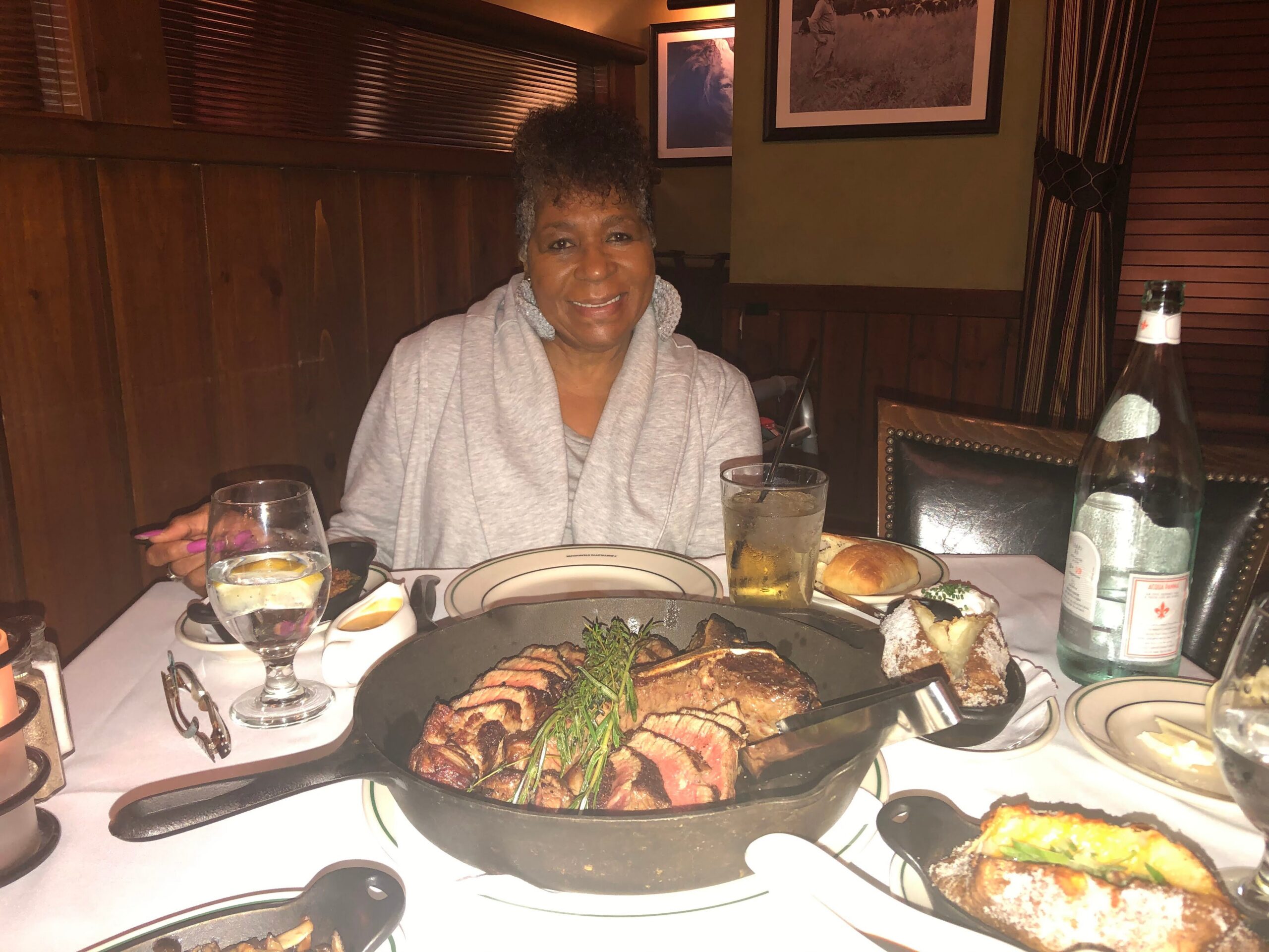 Smiling woman in a grey hoodie seated at a booth with a large steak platter garnished with rosemary in front of her, in a dim restaurant setting with wine bottle and glasses nearby.