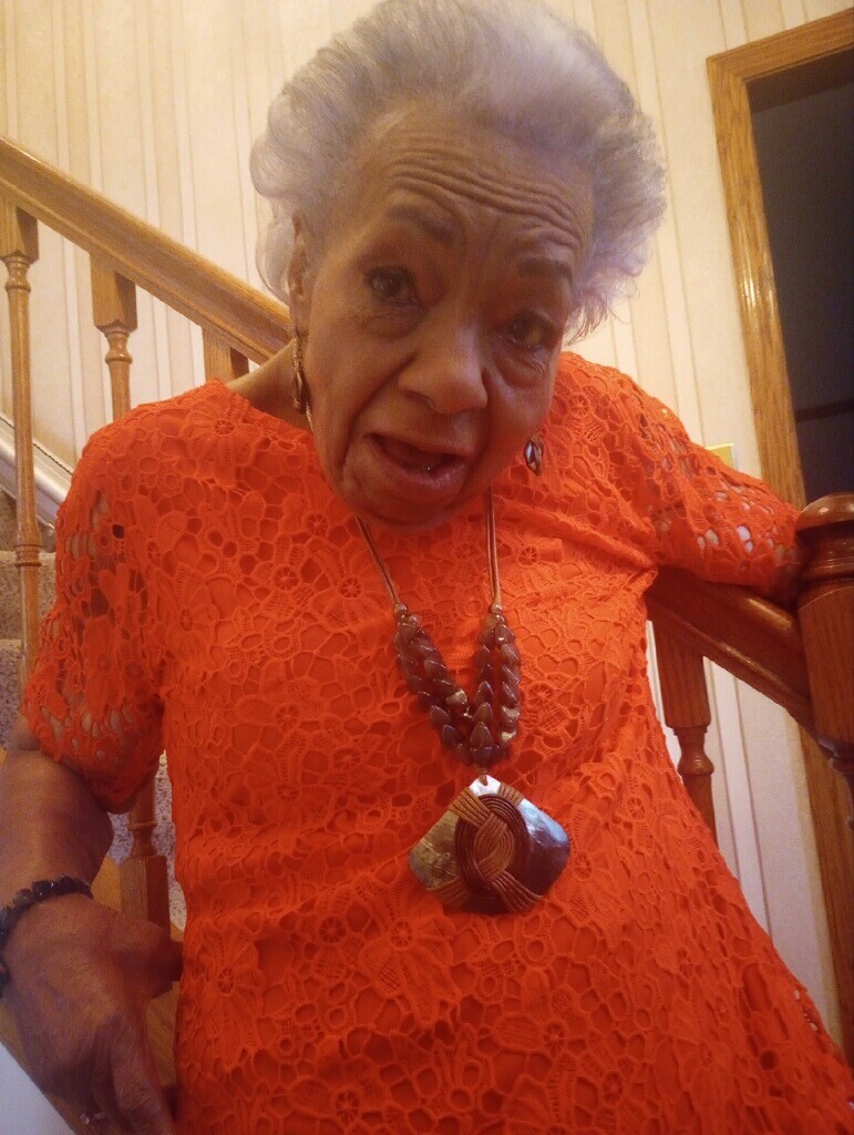 Older woman on a wooden staircase wearing a bright orange lace dress, with bold layered necklace and earrings, looking at the camera.
