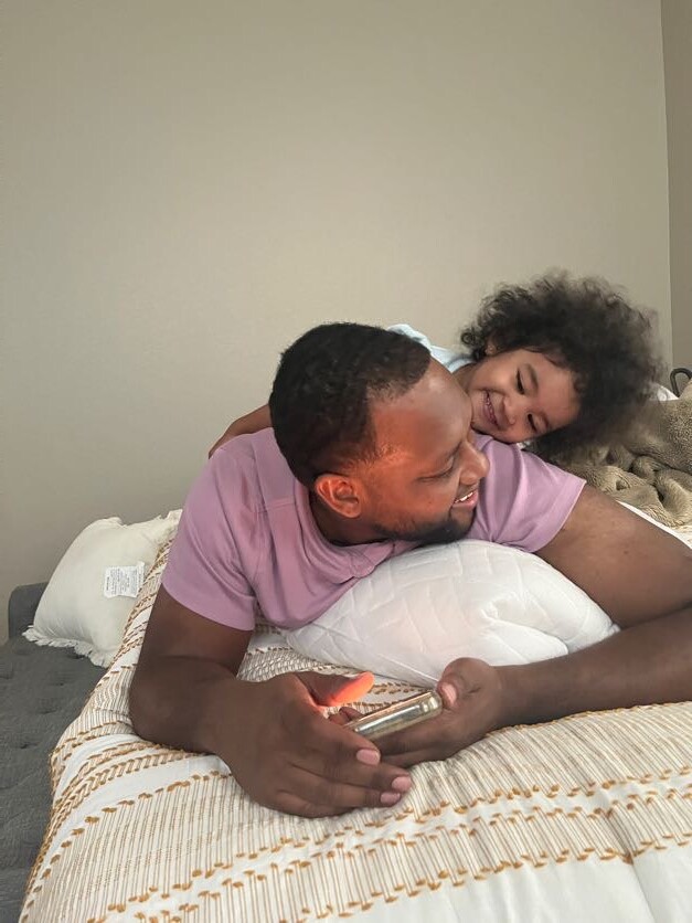 Father and young child cuddle on a bed; the child wraps arms around the man's shoulders as he holds a phone and smiles at them.