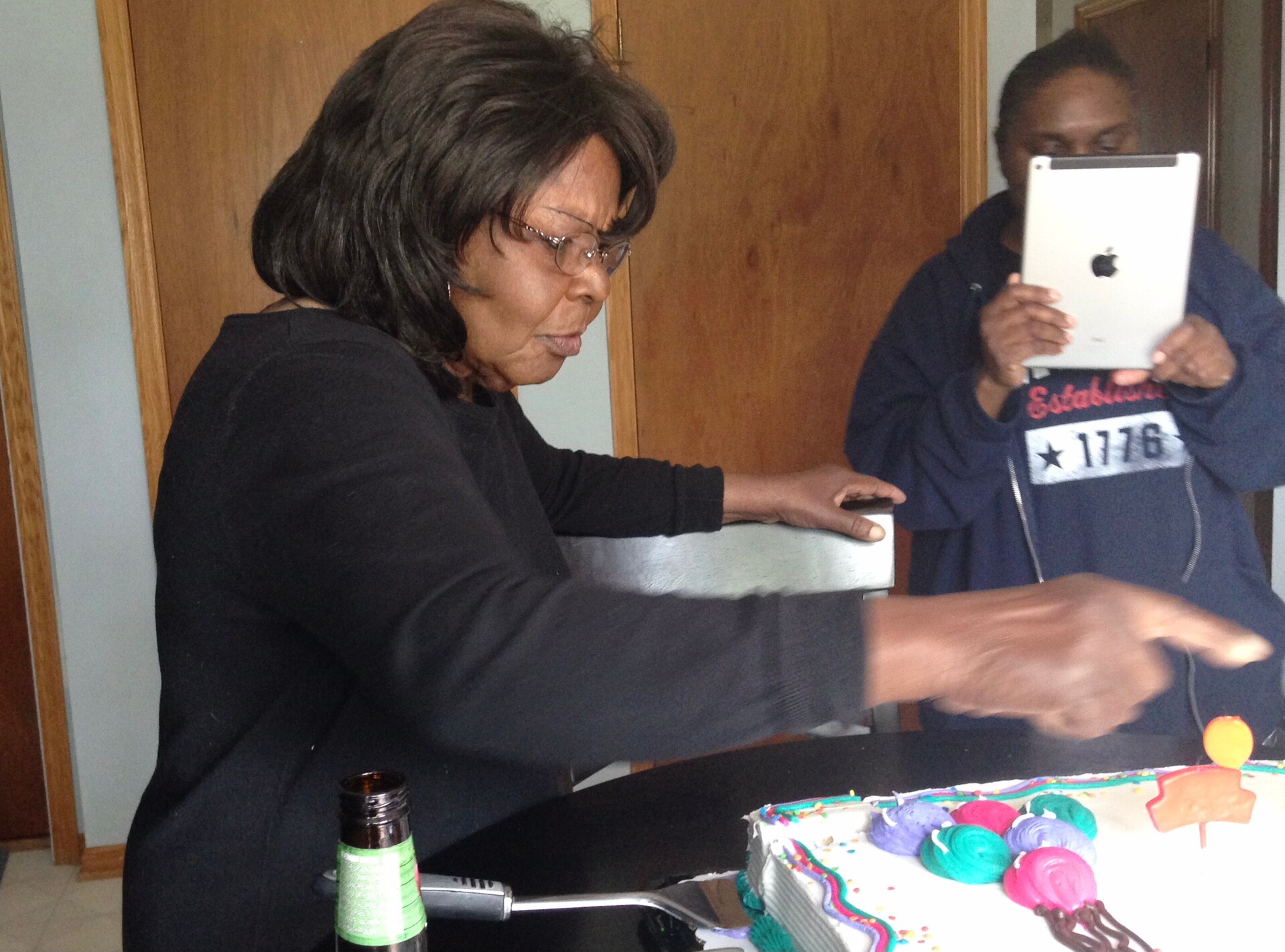 Woman in a black shirt cuts a colorful birthday cake on a kitchen counter while another person holds a tablet nearby.