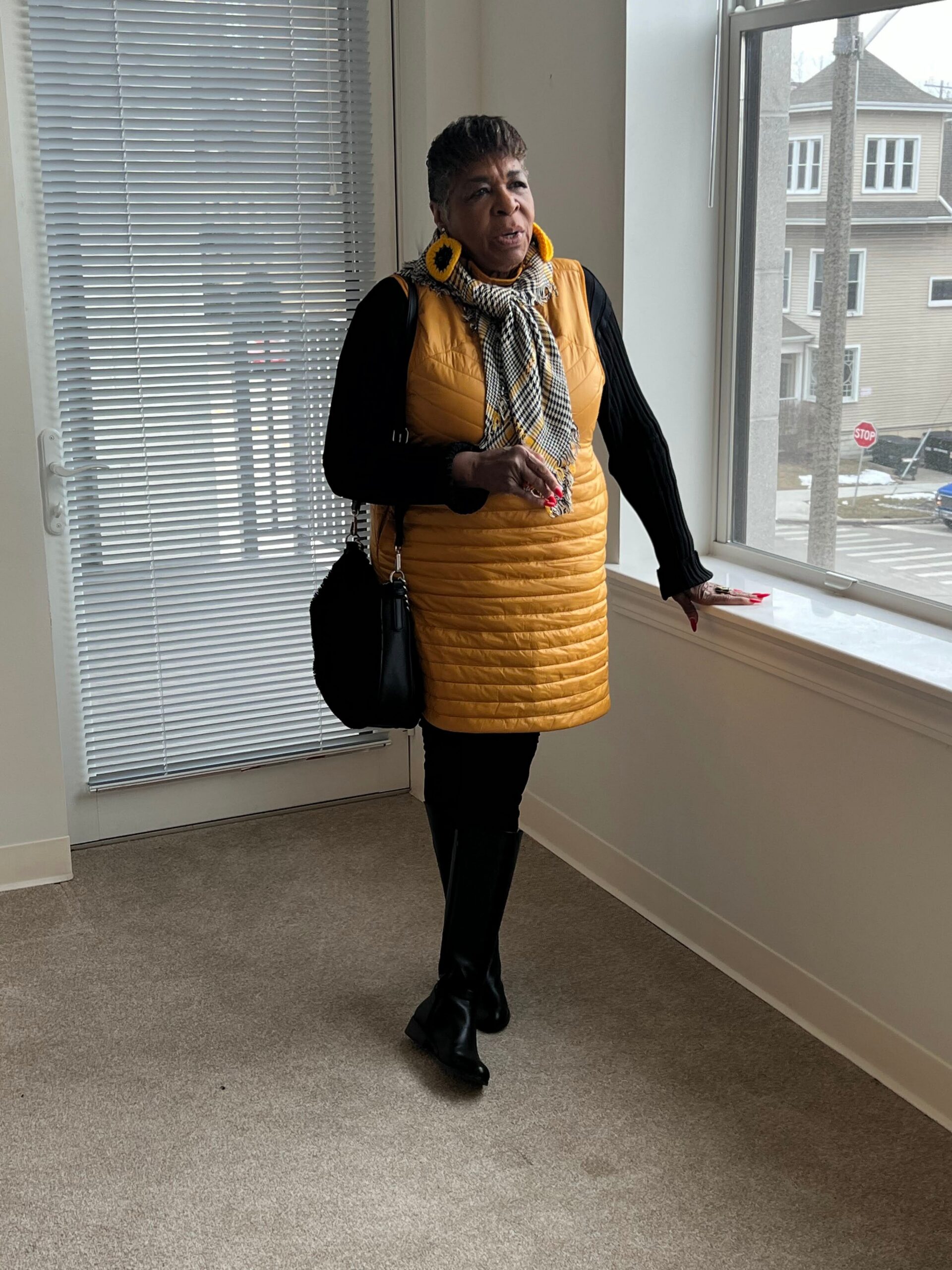 Woman standing indoors by a window in a mustard-yellow quilted dress, black sleeves, plaid scarf, and sunflower earrings, holding a small item near her hand.