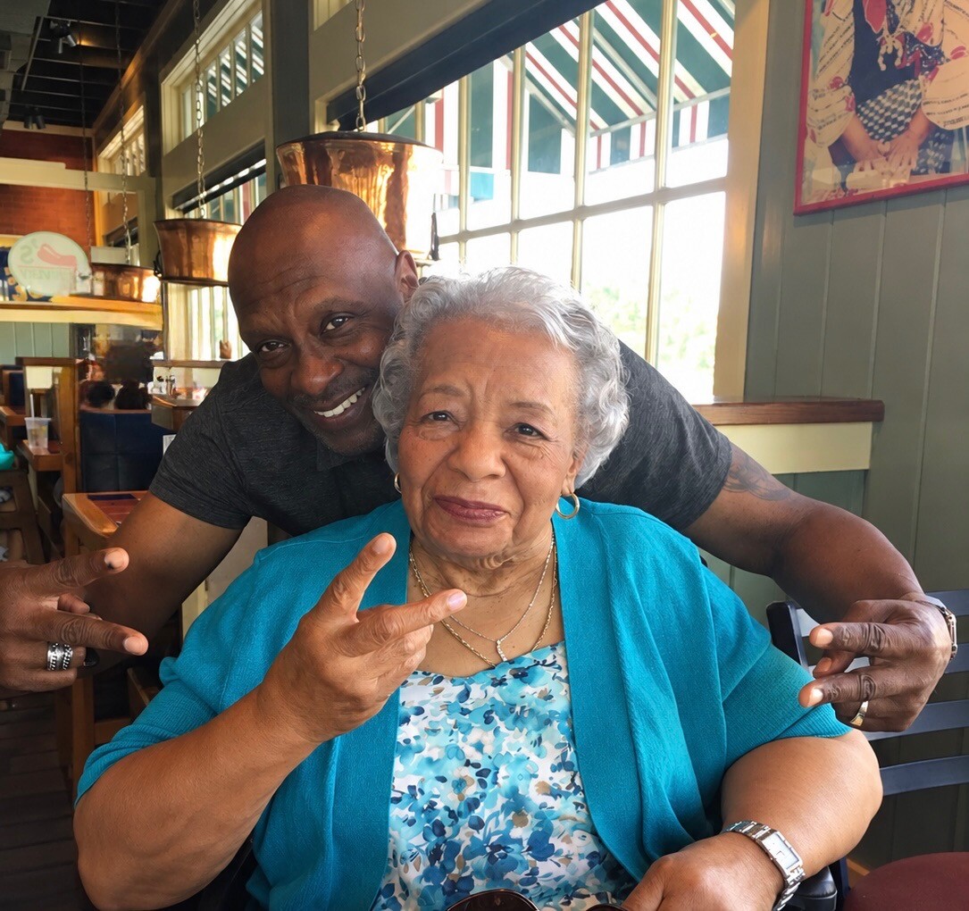 Smiling man behind an elderly woman, both making peace signs at a restaurant table.