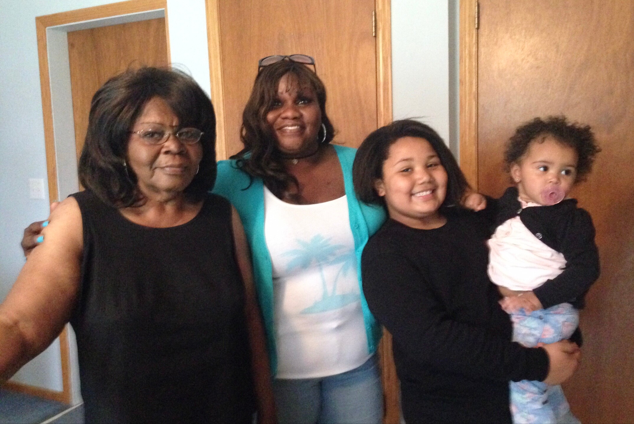 Group of four women with a baby posing together in front of wooden doors; smiling and dressed casually.