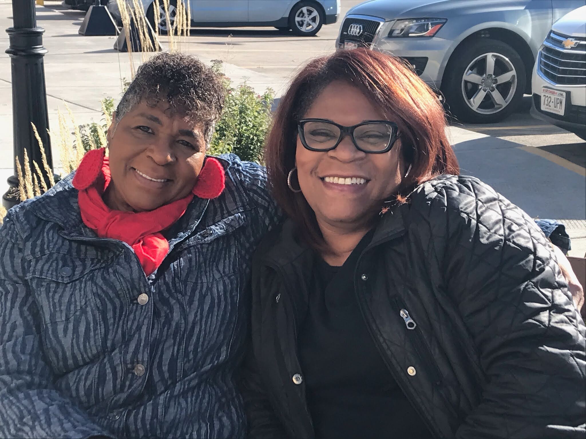 Two women smiling for a selfie in a parking lot, wearing jackets with a blue pattern and a black coat, cars in the background.
