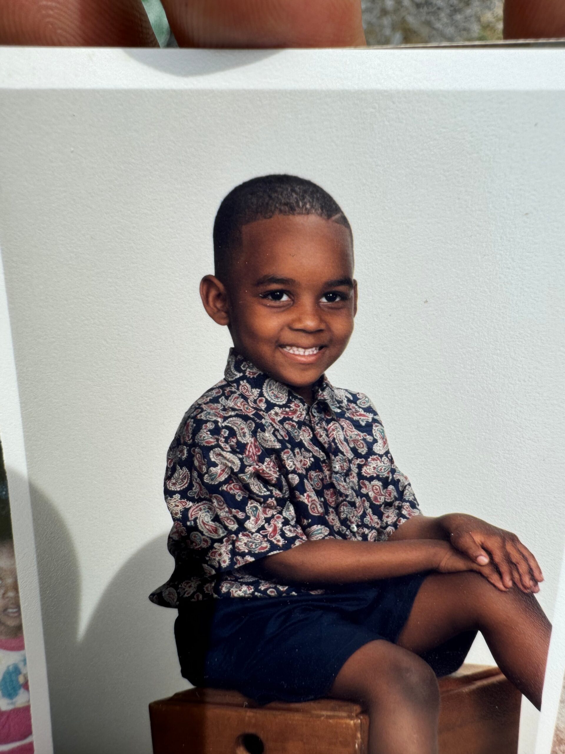 Young boy seated on a wooden stool, smiling, wearing a patterned short-sleeve shirt and dark shorts against a light background.