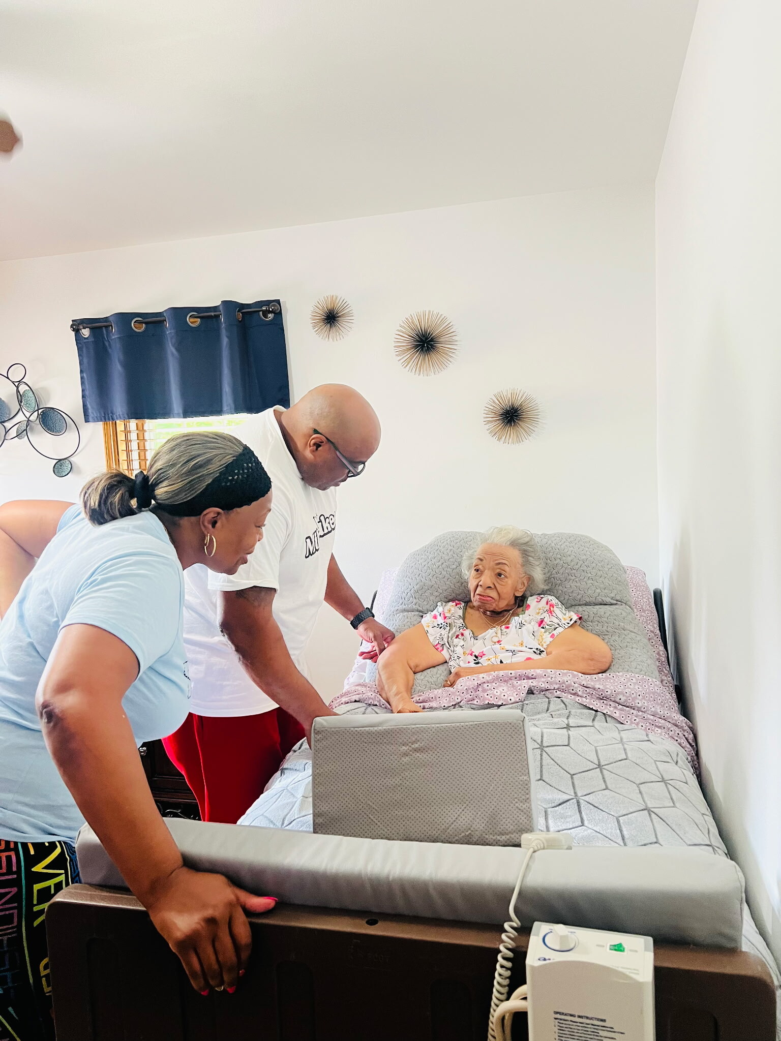 Caregivers attend to an elderly woman resting in a hospital-style bed in a bedroom setting with a medical device nearby
