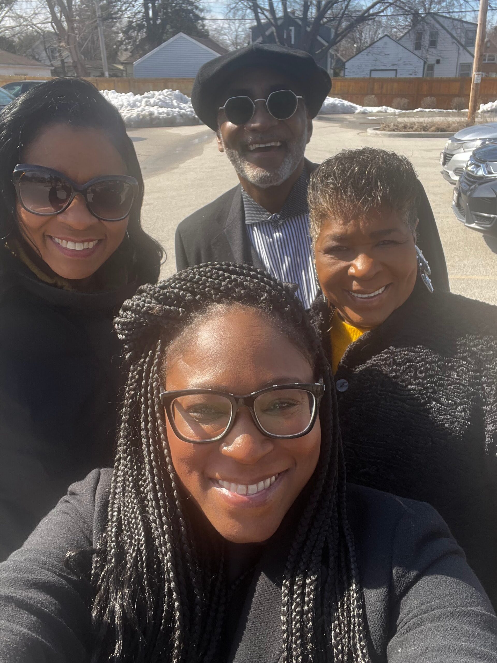 Group selfie of four adults smiling outdoors on a sunny day, with two women wearing sunglasses and a man wearing a hat in the center.