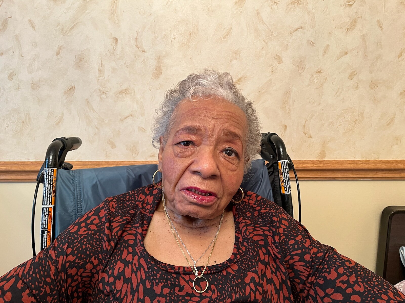 Elderly Black woman in a wheelchair, wearing a red and black patterned top, gold hoop earrings, and a necklace indoors.