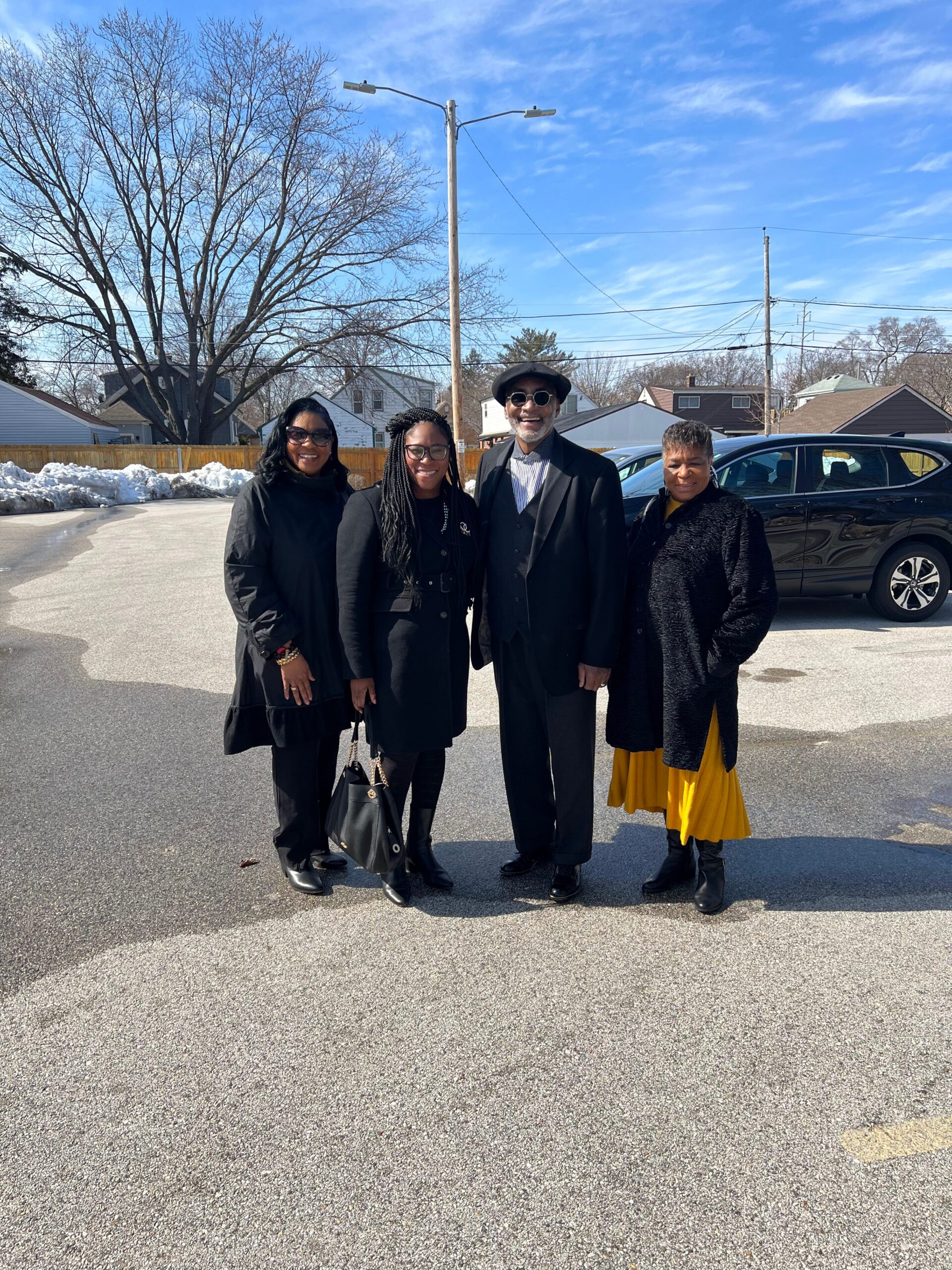 Group of four adults standing together in a parking lot on a sunny winter day, all wearing dark coats; man in a hat and sunglasses in the center-right, woman in a bright yellow skirt on the far right.