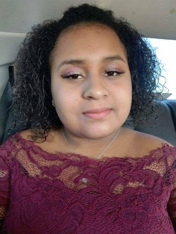 Woman with short curly hair in a car, wearing a maroon lace top and necklace, looking at the camera.