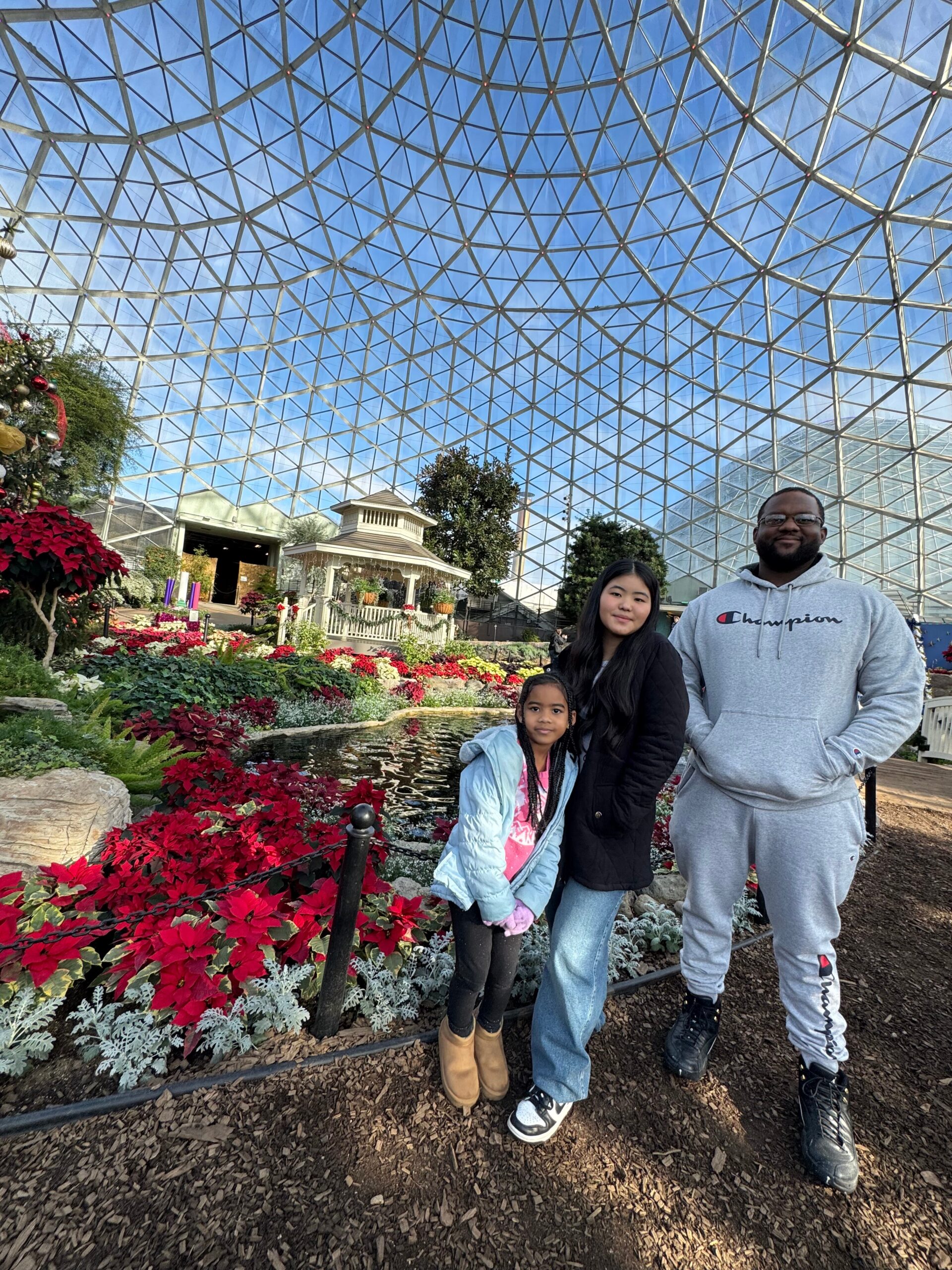 Three people pose together in a glass-domed conservatory with red poinsettias and a small gazebo in the background.