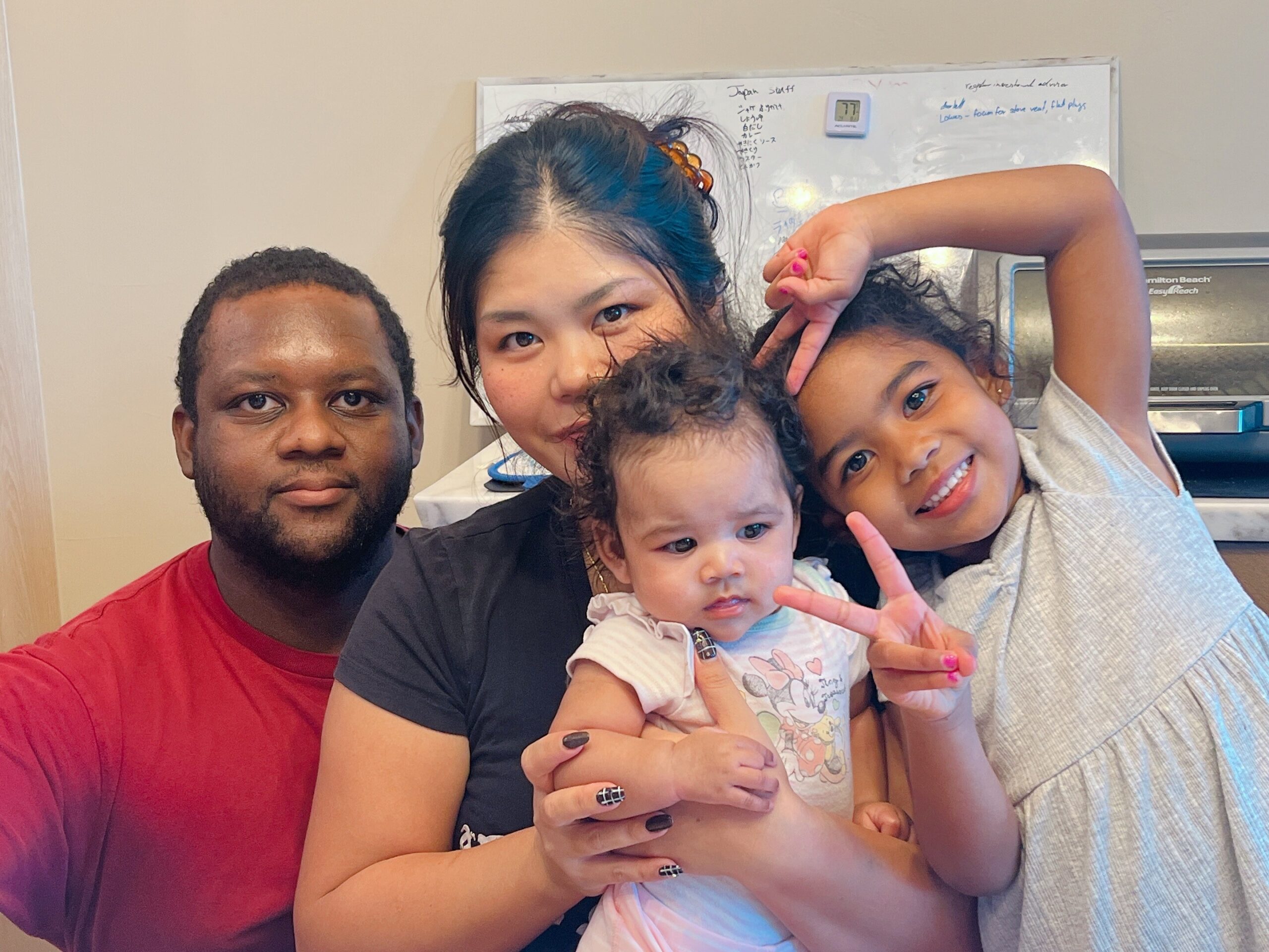 A family of four with a baby posing for a selfie indoors, with the father on the left and the mother in the center flanked by two children.