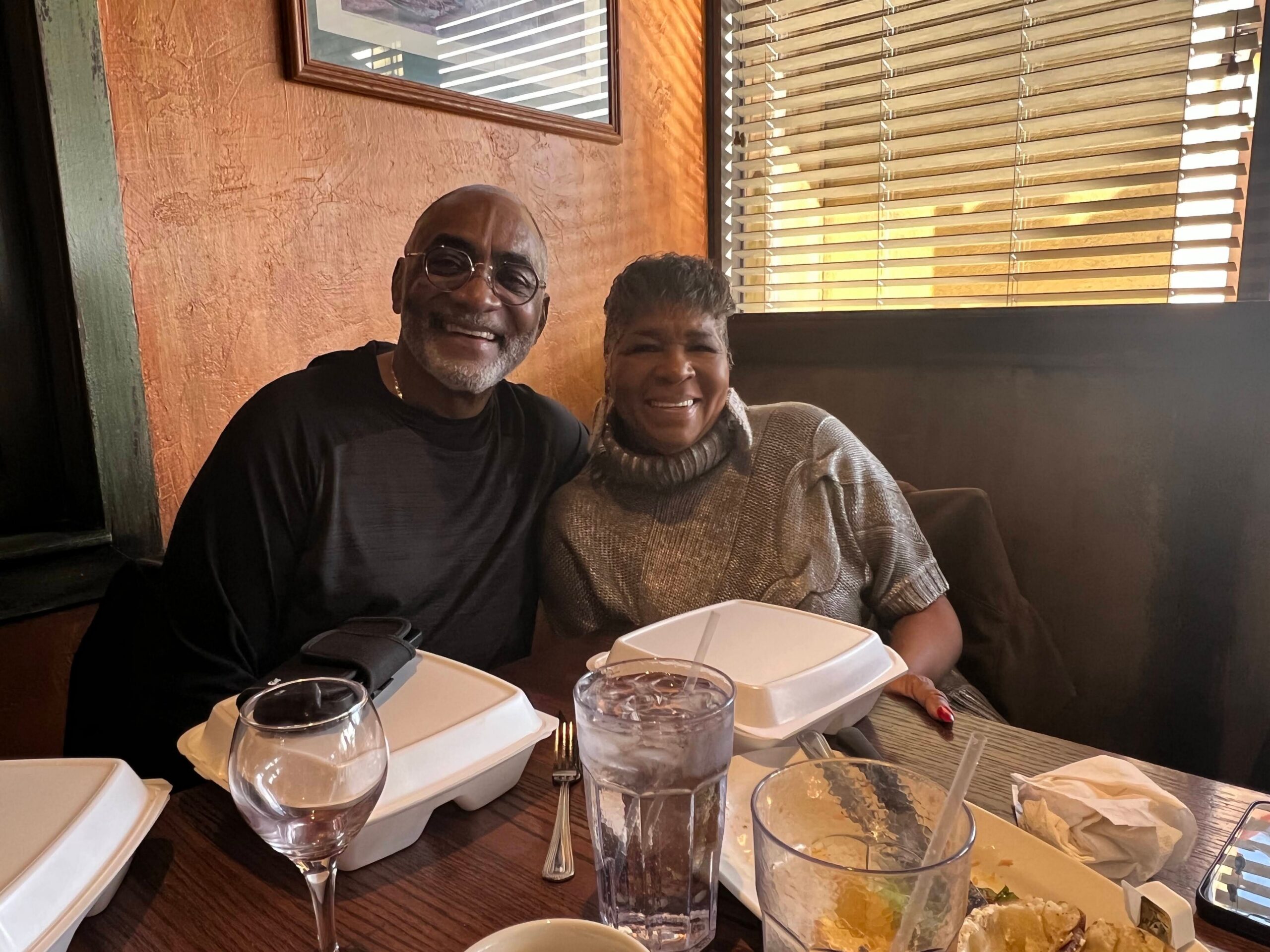 Smiling couple sitting at a restaurant table with takeout boxes and drinks in front of them indoors.