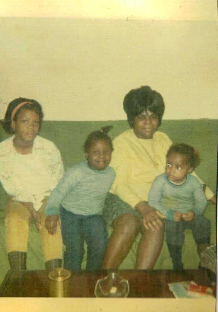 Four people sit on a couch: two adults in the center and two young children on either side, posing for a family photo in a living room.