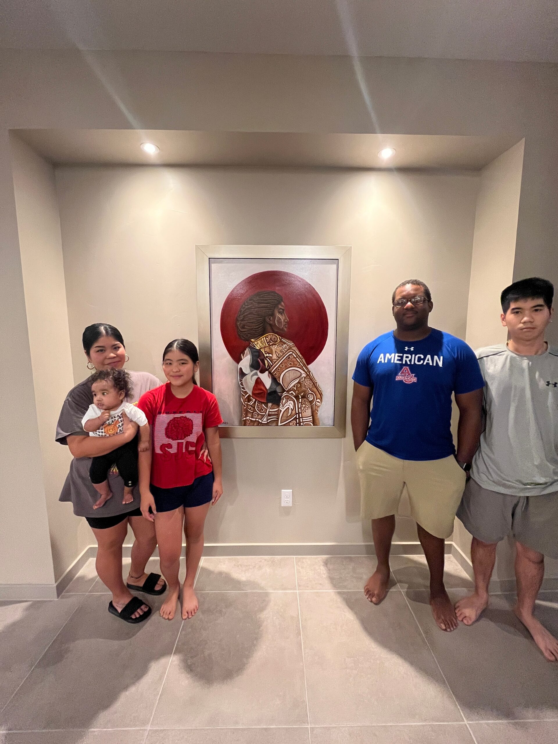 A group of six people posing in a hallway in front of a framed painting; two children, four adults, some barefoot, casual clothing under ceiling spotlights.