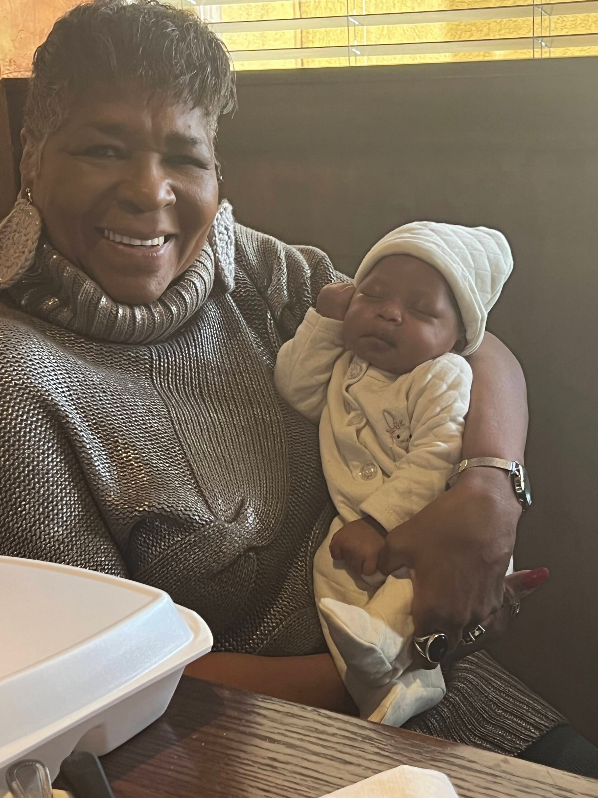 Smiling woman in a metallic knit sweater holding a sleeping newborn wrapped in a white blanket and hat in a restaurant booth.