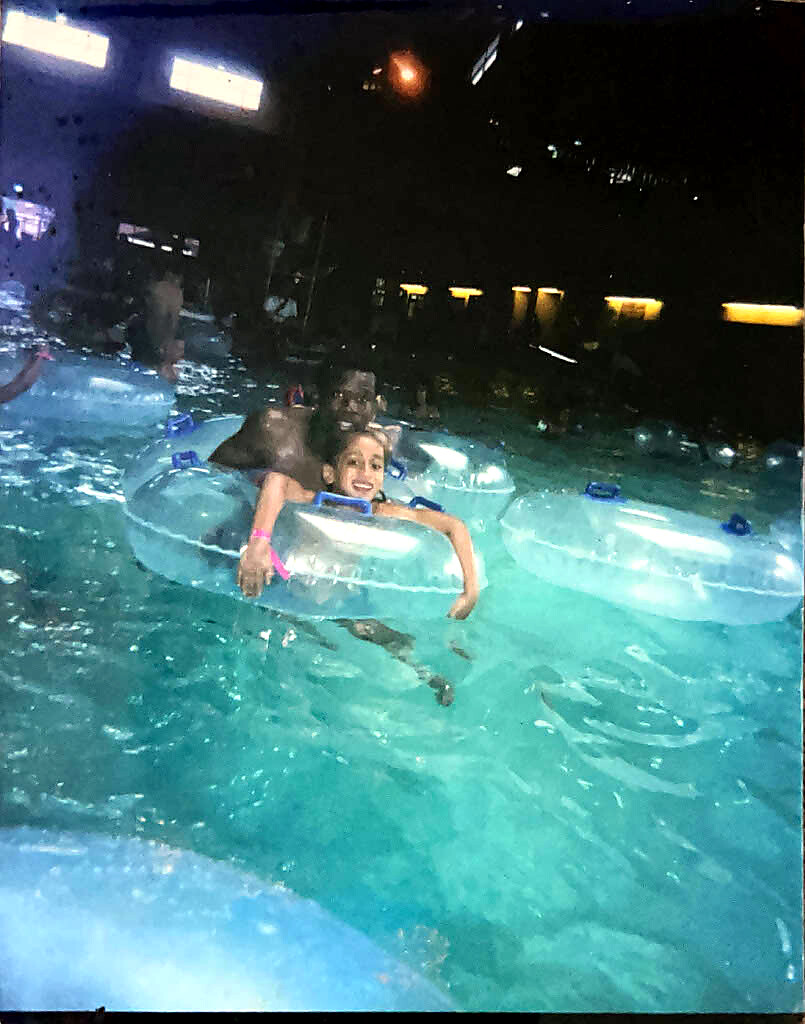 Two people in clear inflatable tubes float in a turquoise pool at night, smiling at the camera.