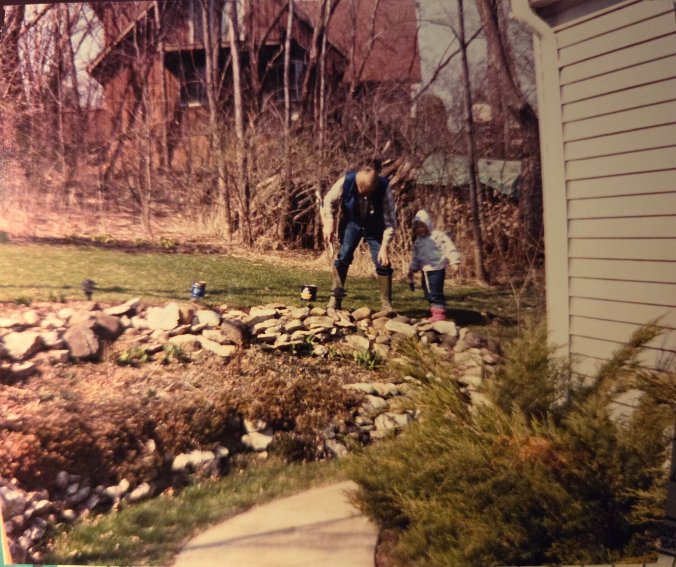 A man and a child bend over in a yard by a rocky edge, with a beige house wall on the right and leafless trees in the background.