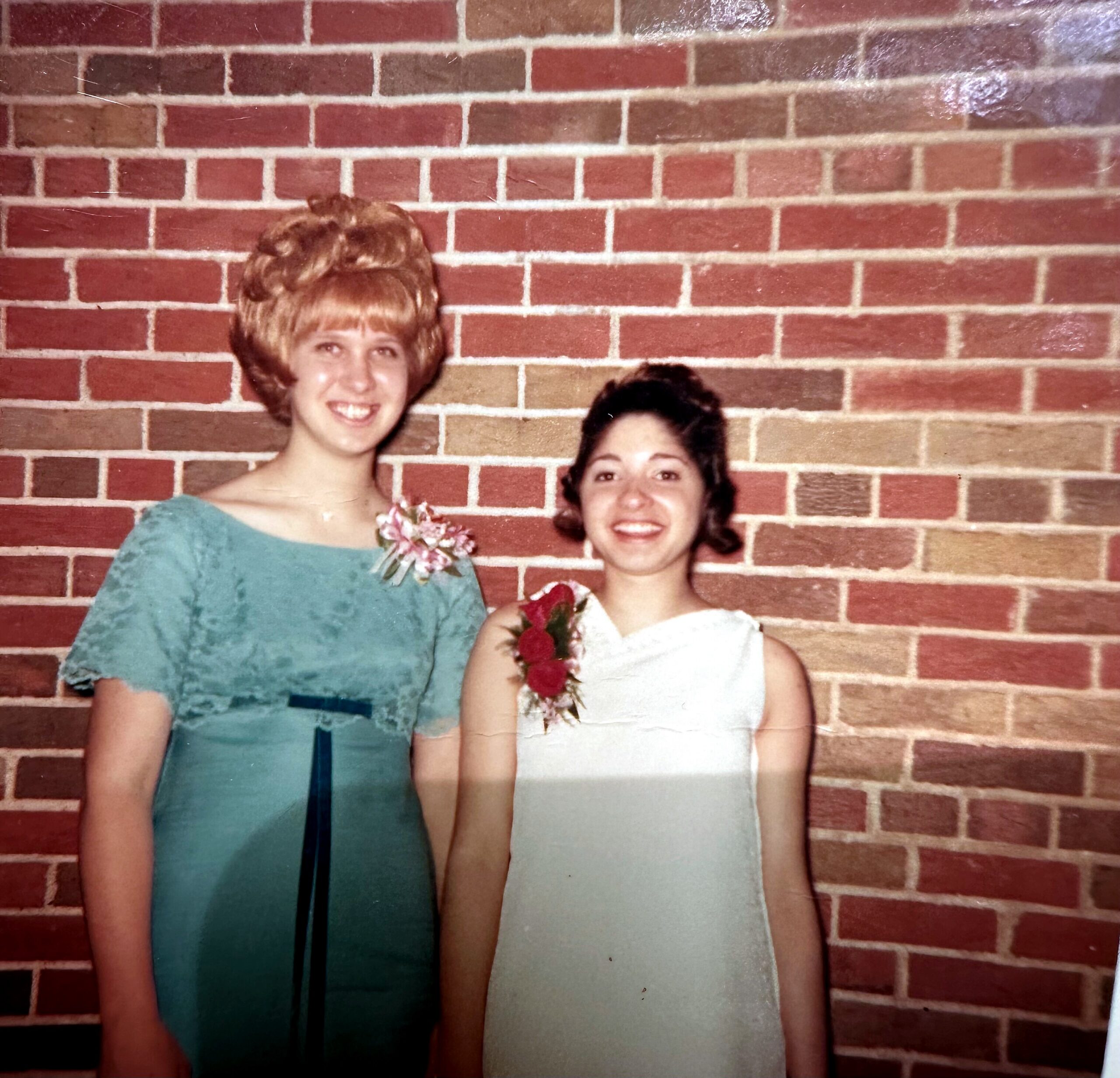 Two women pose together in front of a red brick wall; the left woman wears a teal dress with a corsage, the right woman wears a white dress with a floral accessory.
