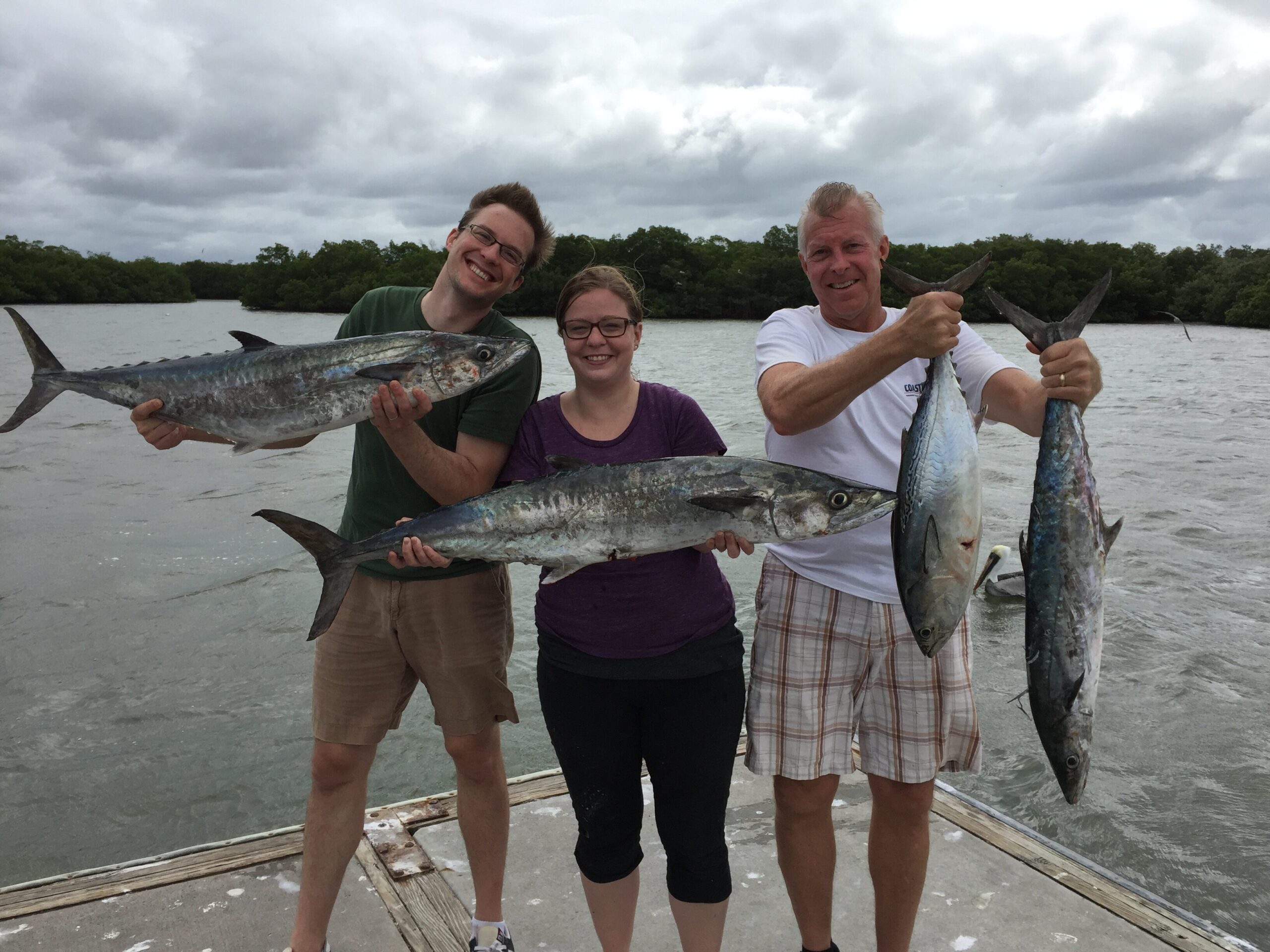 Three people stand on a dock, each holding a large fish, with a cloudy sky and water in the background.