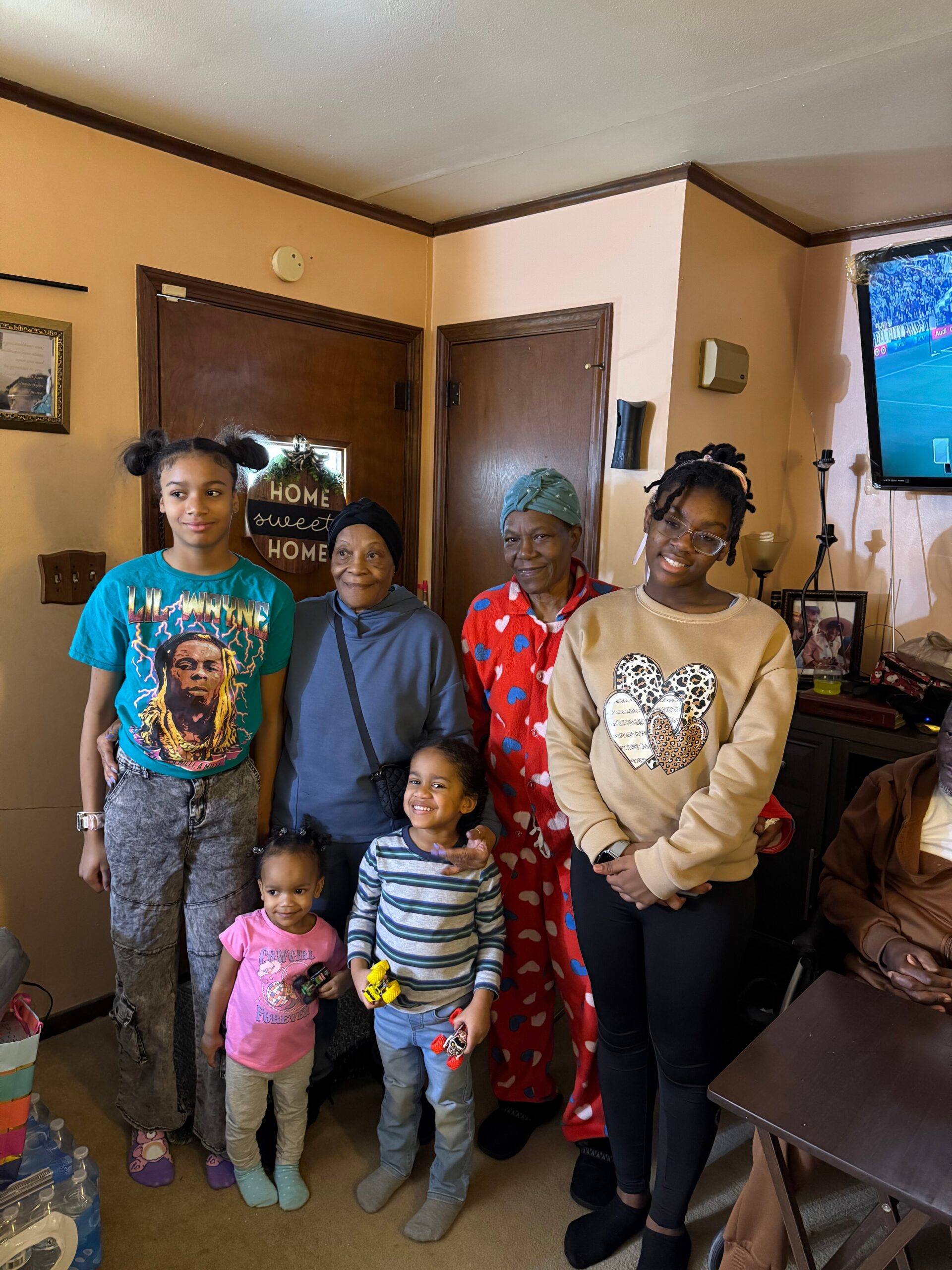 Group of people posing for a photo in a living room; a TV shows a sports game in the background. (Multigenerational family)