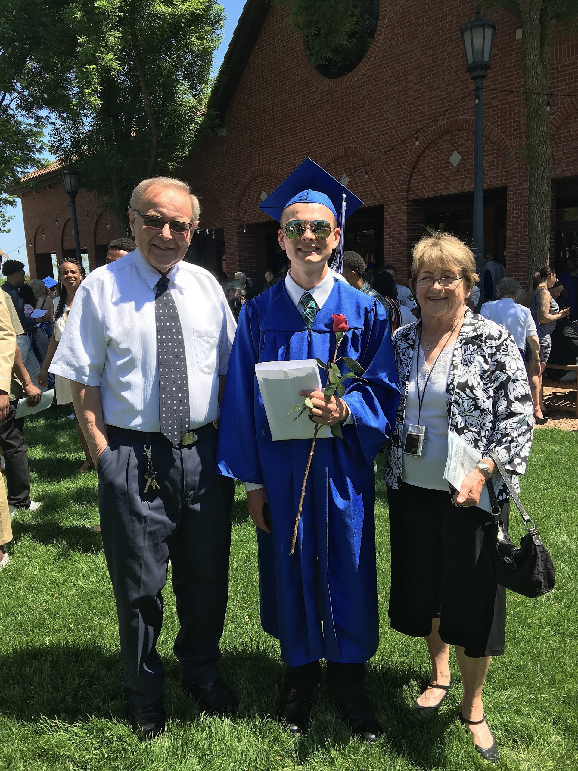 Graduate in blue cap and gown standing between an older man in a white shirt and a woman in a floral jacket outdoors on a sunny day by a brick building.