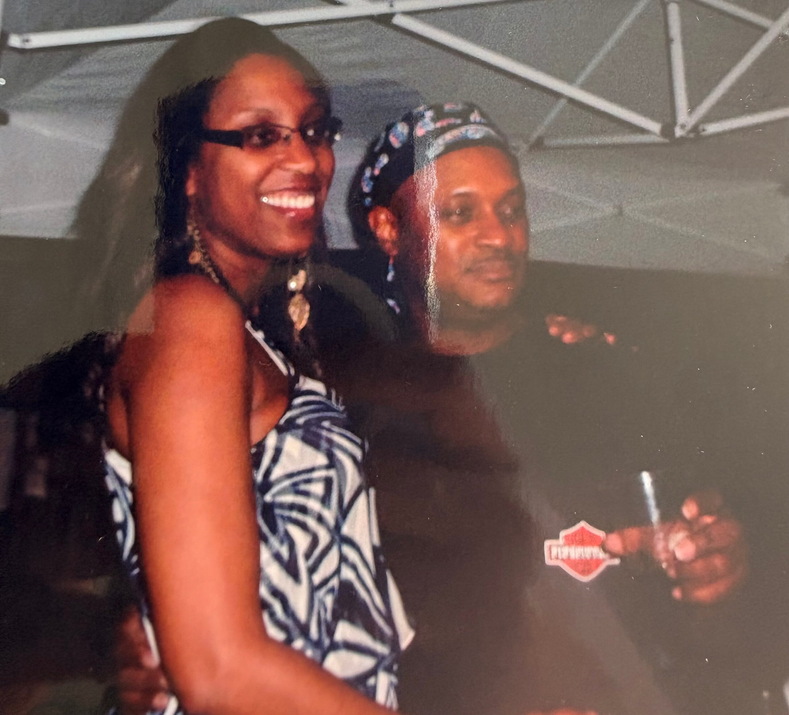 Two friends posing for a photo, smiling; left person wears glasses and a striped top, right person wears a patterned headscarf.