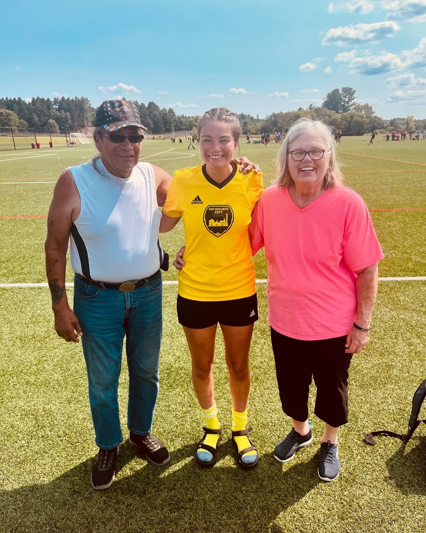 Three people posing on a sunny soccer field: man in a blue sleeveless shirt left, girl in a yellow Milwaukee City jersey center, and woman in a pink shirt right.