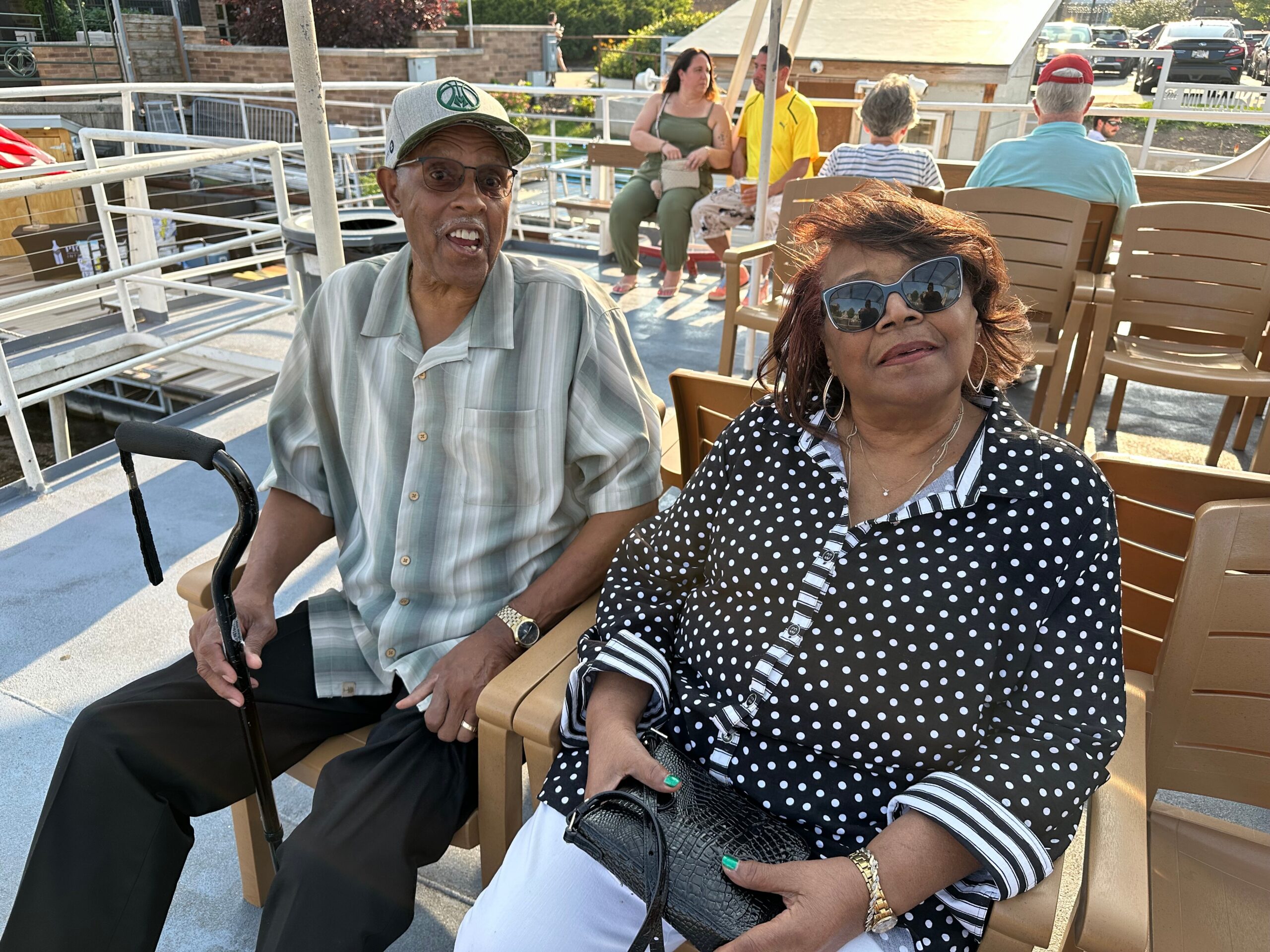 Two older adults sit together on a sunny outdoor boat deck; man with a cane and cap sits beside a woman in a black-and-white polka dot blouse wearing large sunglasses. In the background, other passengers chat and lounge on chairs.