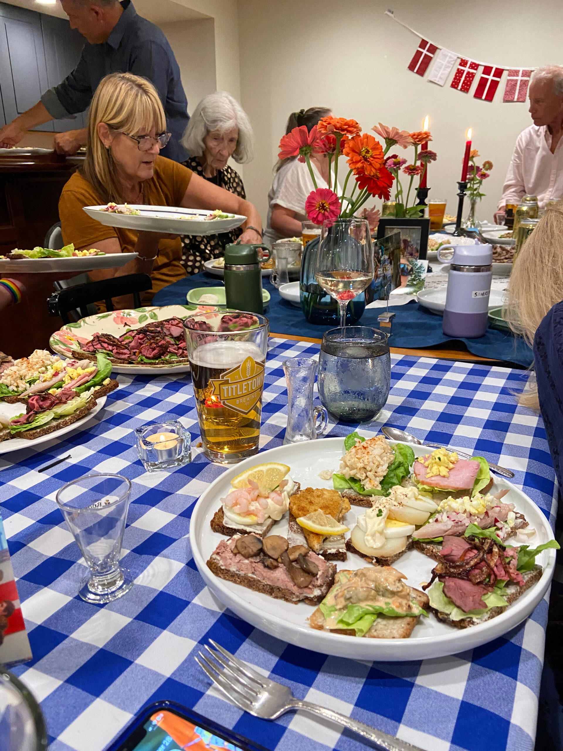 A long table with blue-and-white checkered cloth, plates of open-faced sandwiches, and a centerpiece of orange flowers; people chat in the background as food is served.