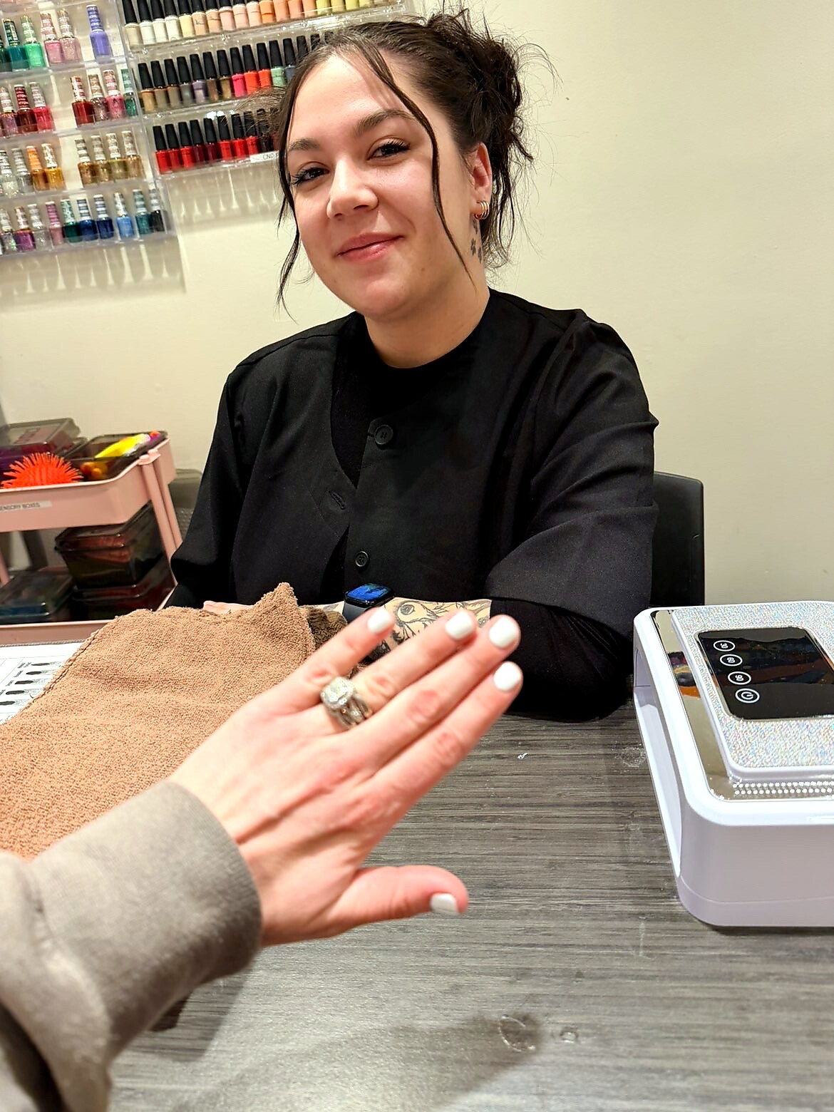 Nail technician in black uniform smiles at a manicure counter as a client shows a white-manicured hand with rings toward the camera; nail polish shelves in the background.