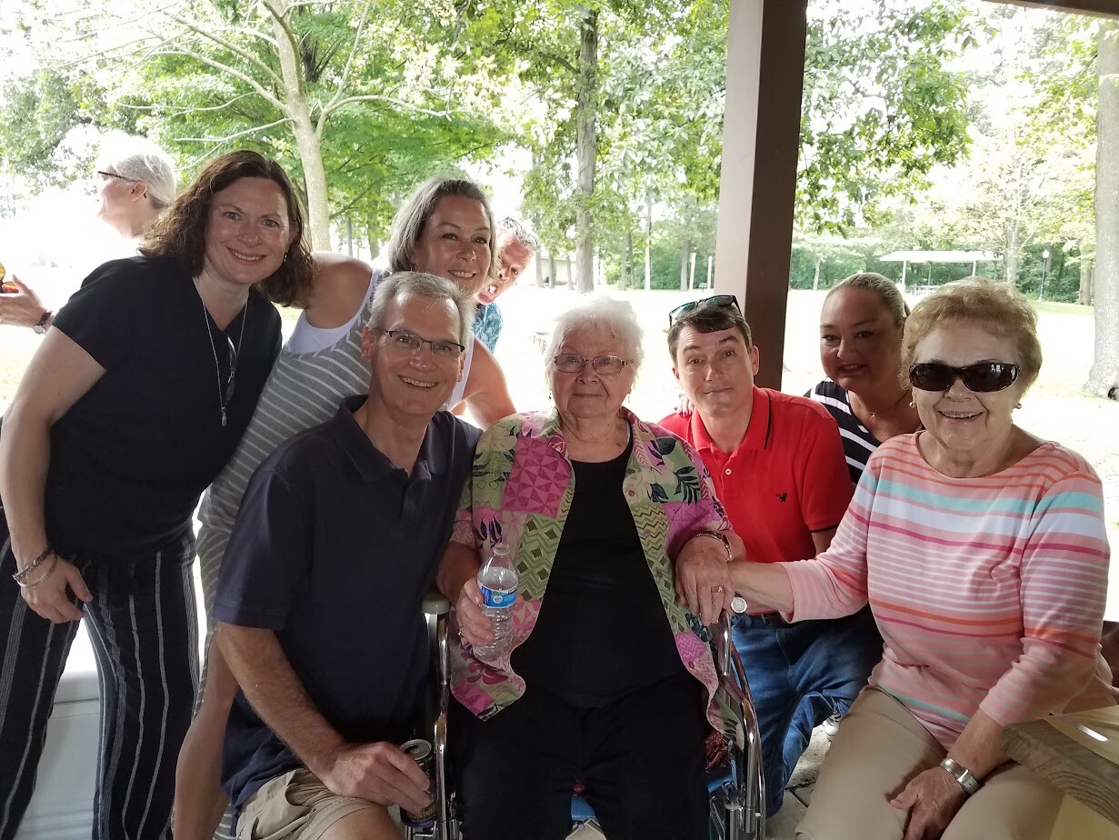 Group of eight people posing under a park pavilion, with an elderly woman in a wheelchair at the center, smiling at the camera.