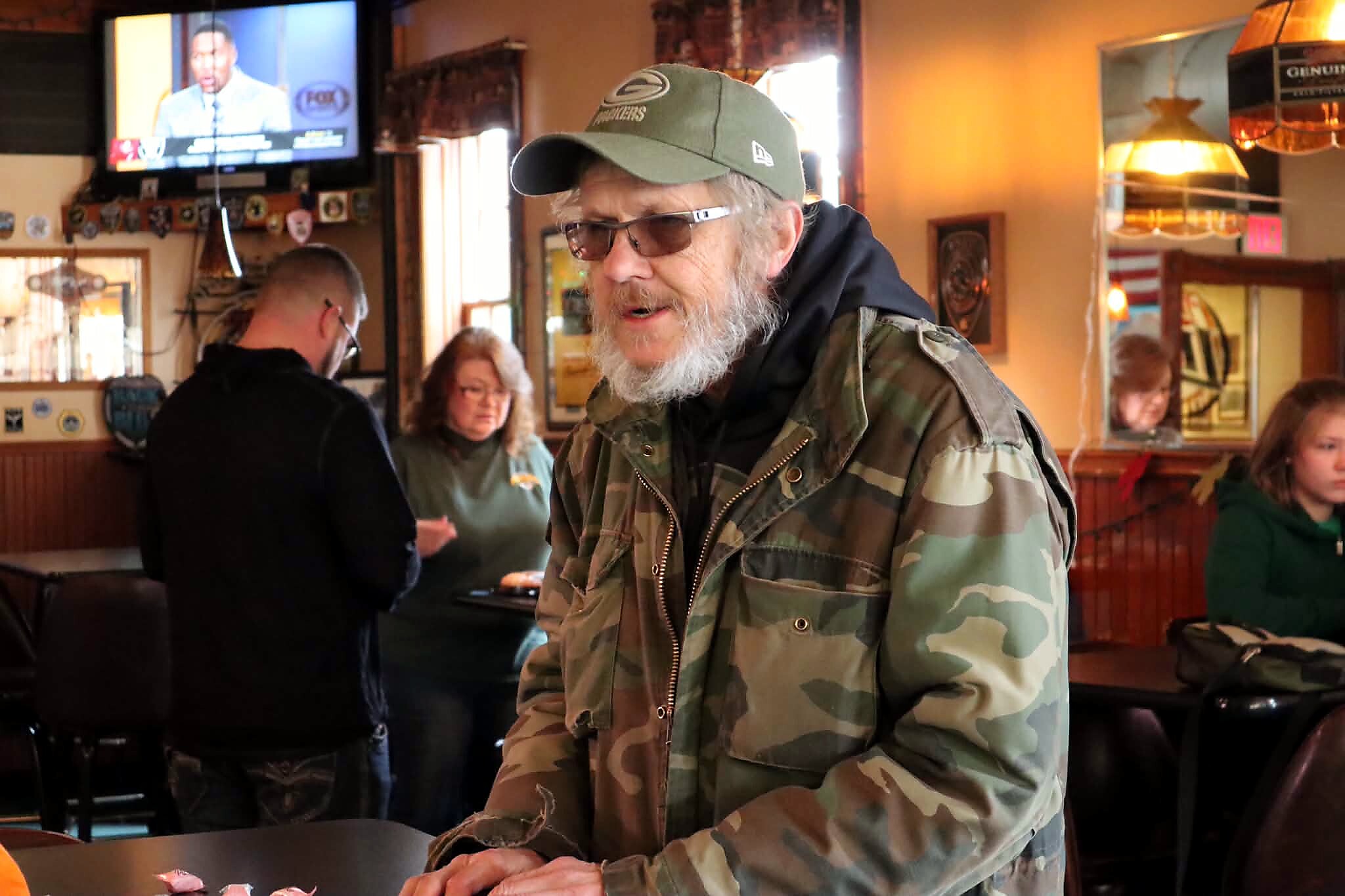 Bearded man in camouflage jacket and green cap standing at a bar indoors.