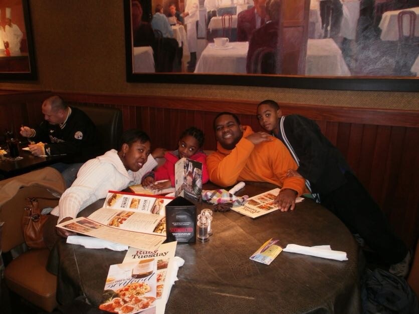 Five friends gathered around a round restaurant table, looking at menus and smiling at the camera.