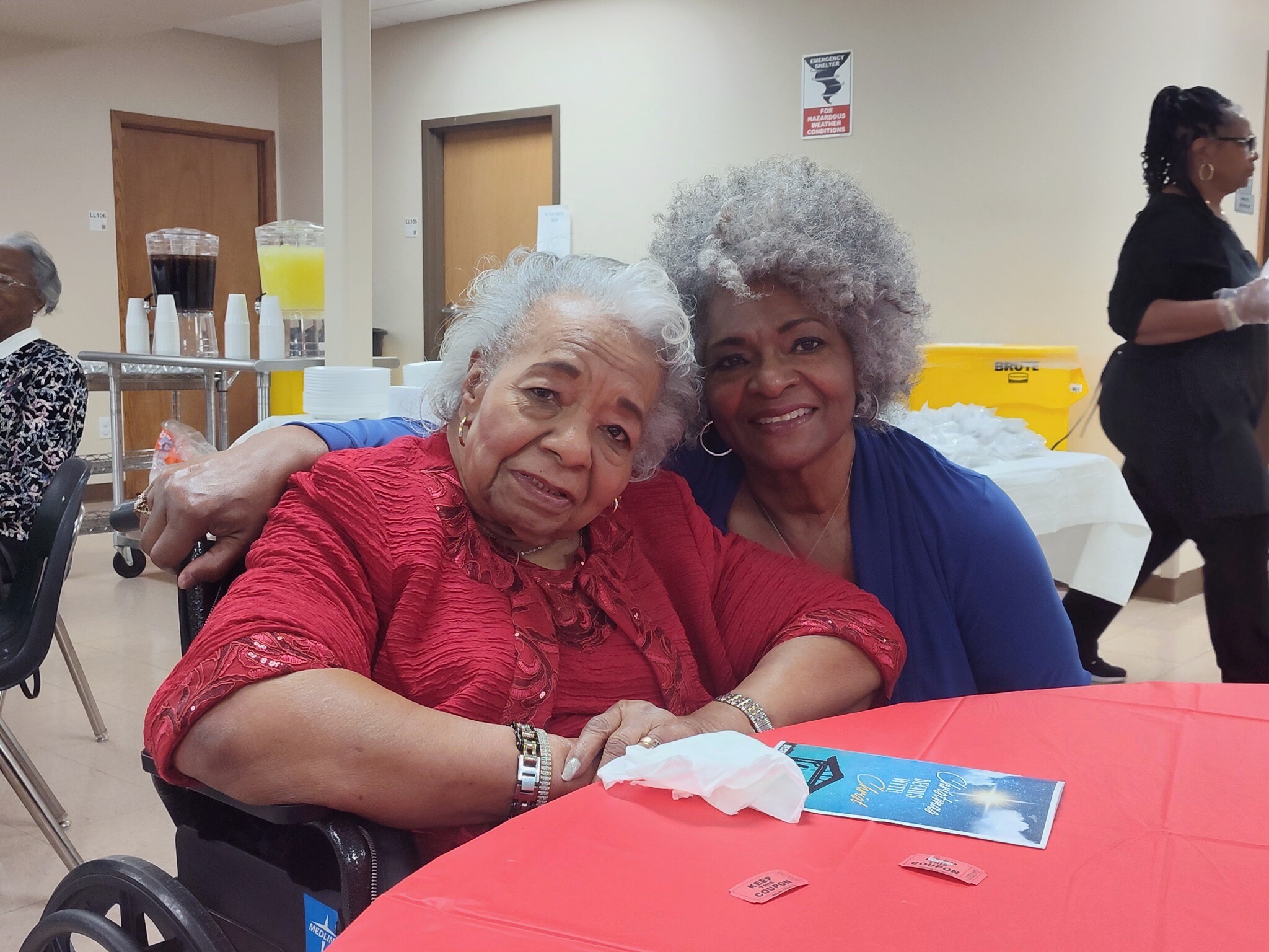 Two smiling women pose for a selfie at a table in a community center, one in a red blouse, the other in blue, with a wheelchair nearby and juice dispensers in the background.