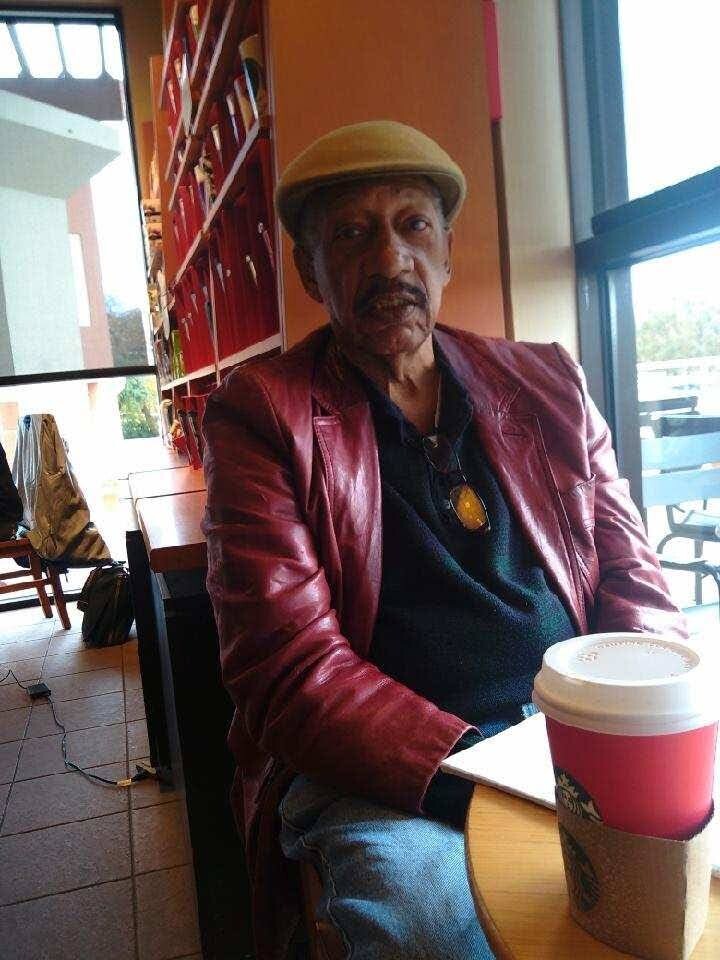 Older man in a tan hat and red leather jacket sits at a cafe table with a takeaway coffee cup in the foreground.