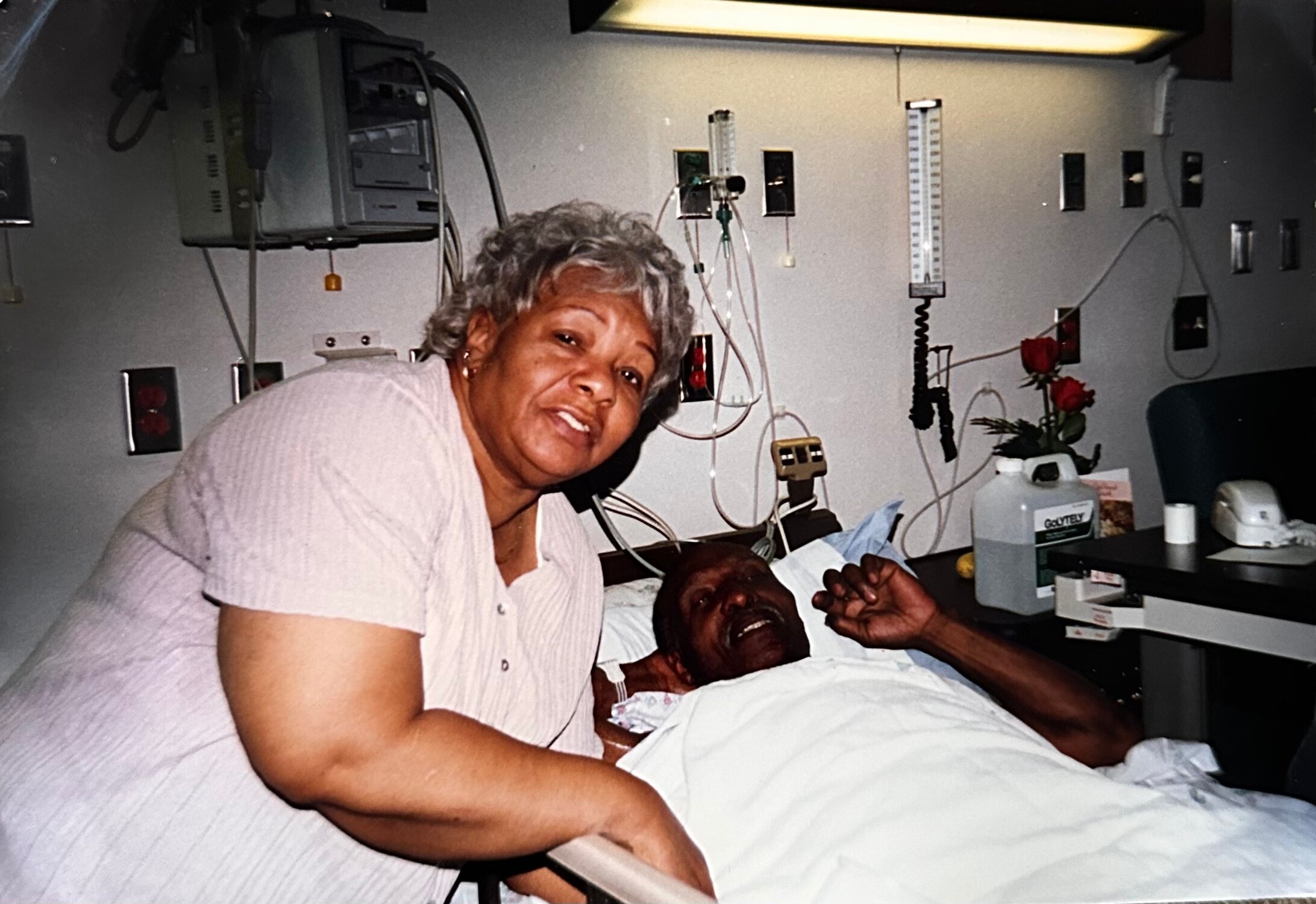 Older woman leaning over a patient in a hospital bed, smiling, with medical equipment on the wall behind them.