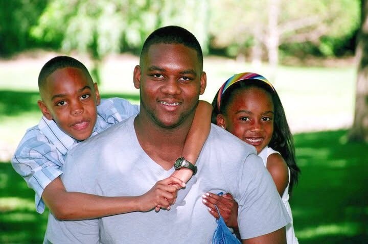 Smiling man in a gray shirt with two children hugging him from behind in a sunny park.