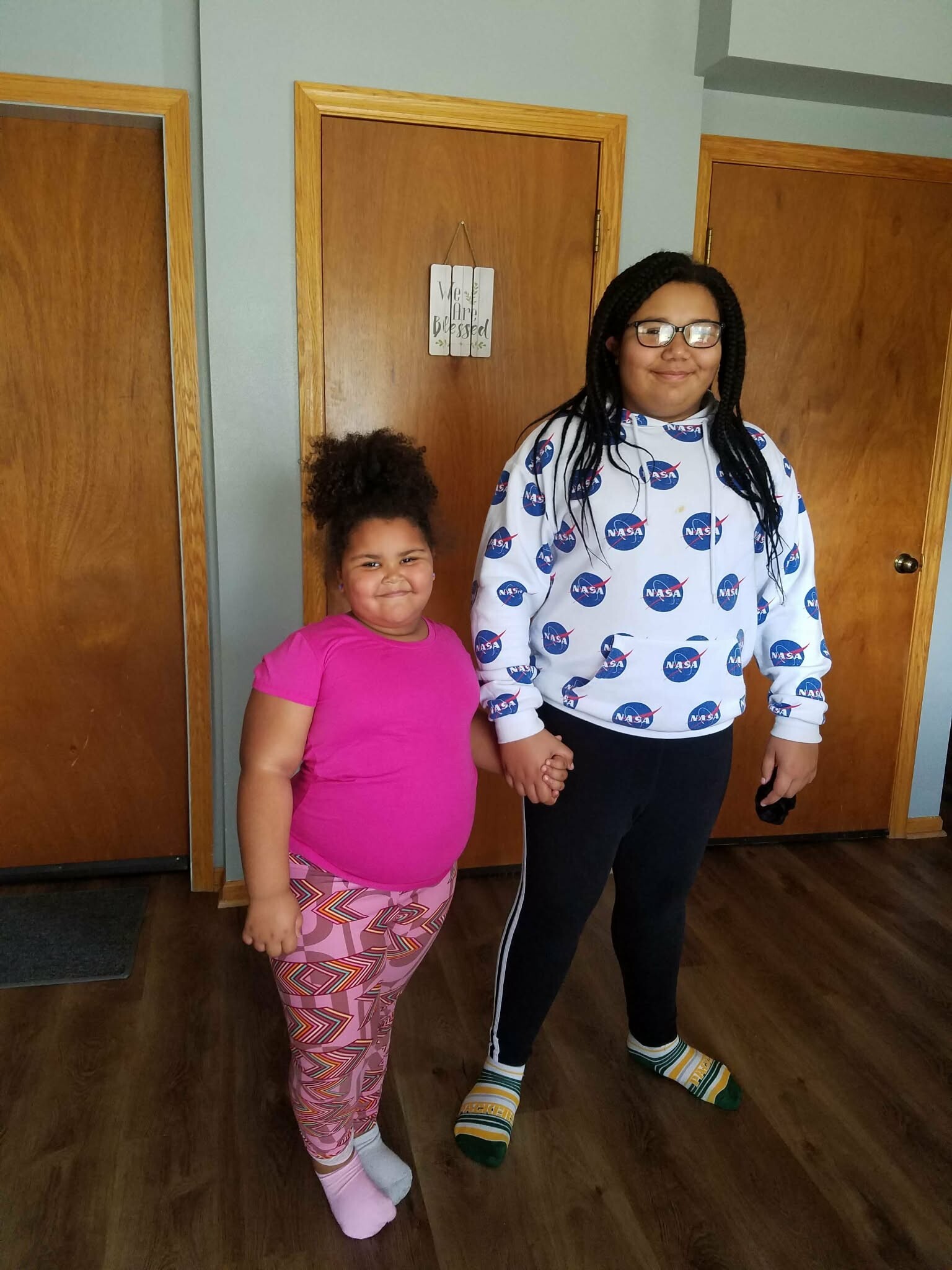 Two girls stand indoors, holding hands; younger girl in a pink shirt and patterned leggings, older girl in a NASA hoodie with striped socks.