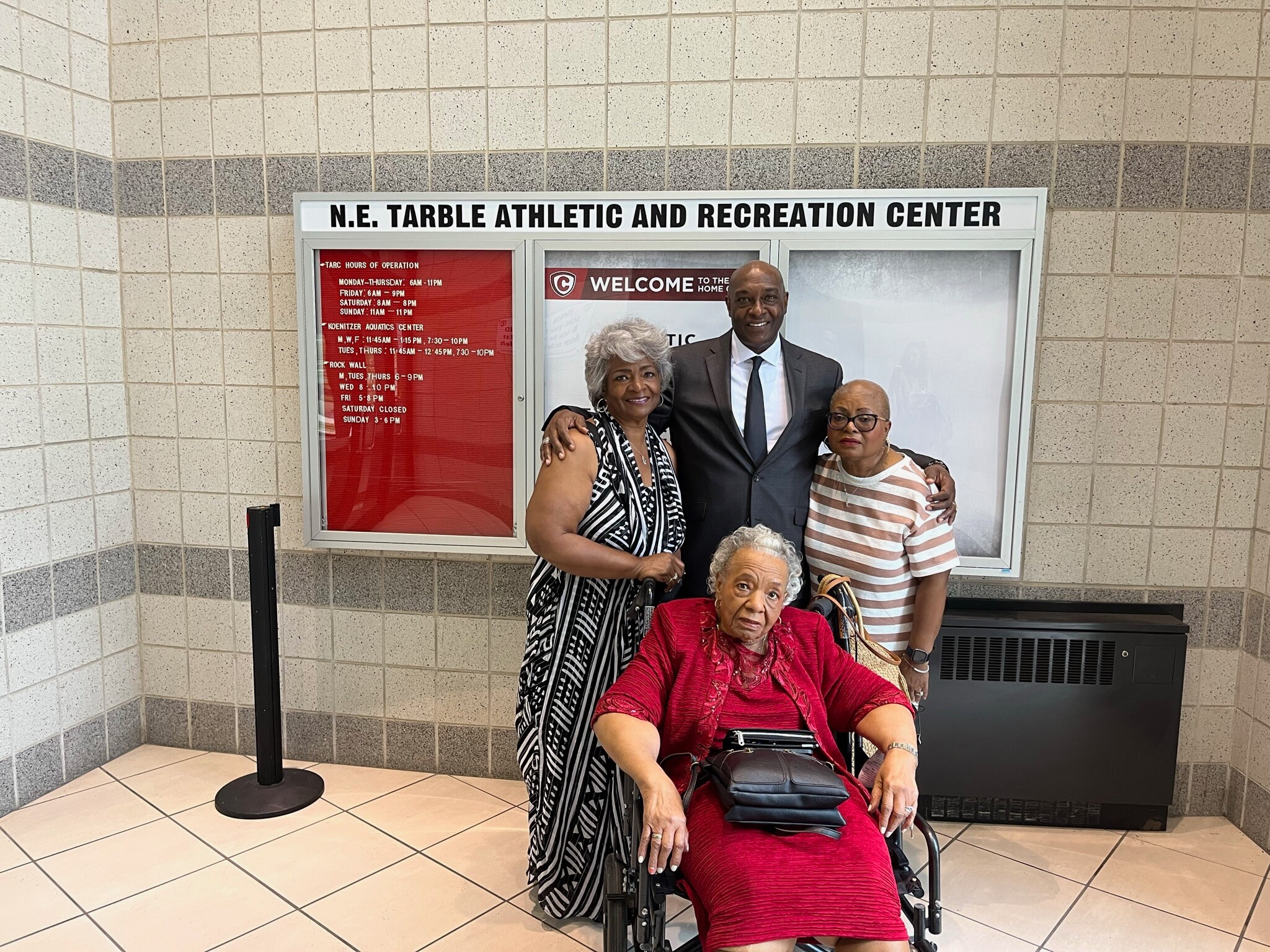 Group of four adults posing together in a tiled lobby, with a woman in a red dress in a wheelchair in front and a sign for N.E. Tarble Athletic and Recreation Center behind them.