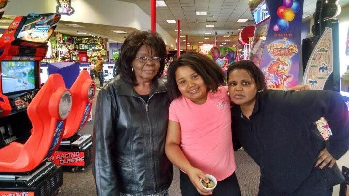 Three women pose and smile in an arcade, with colorful game machines and balloons in the background.