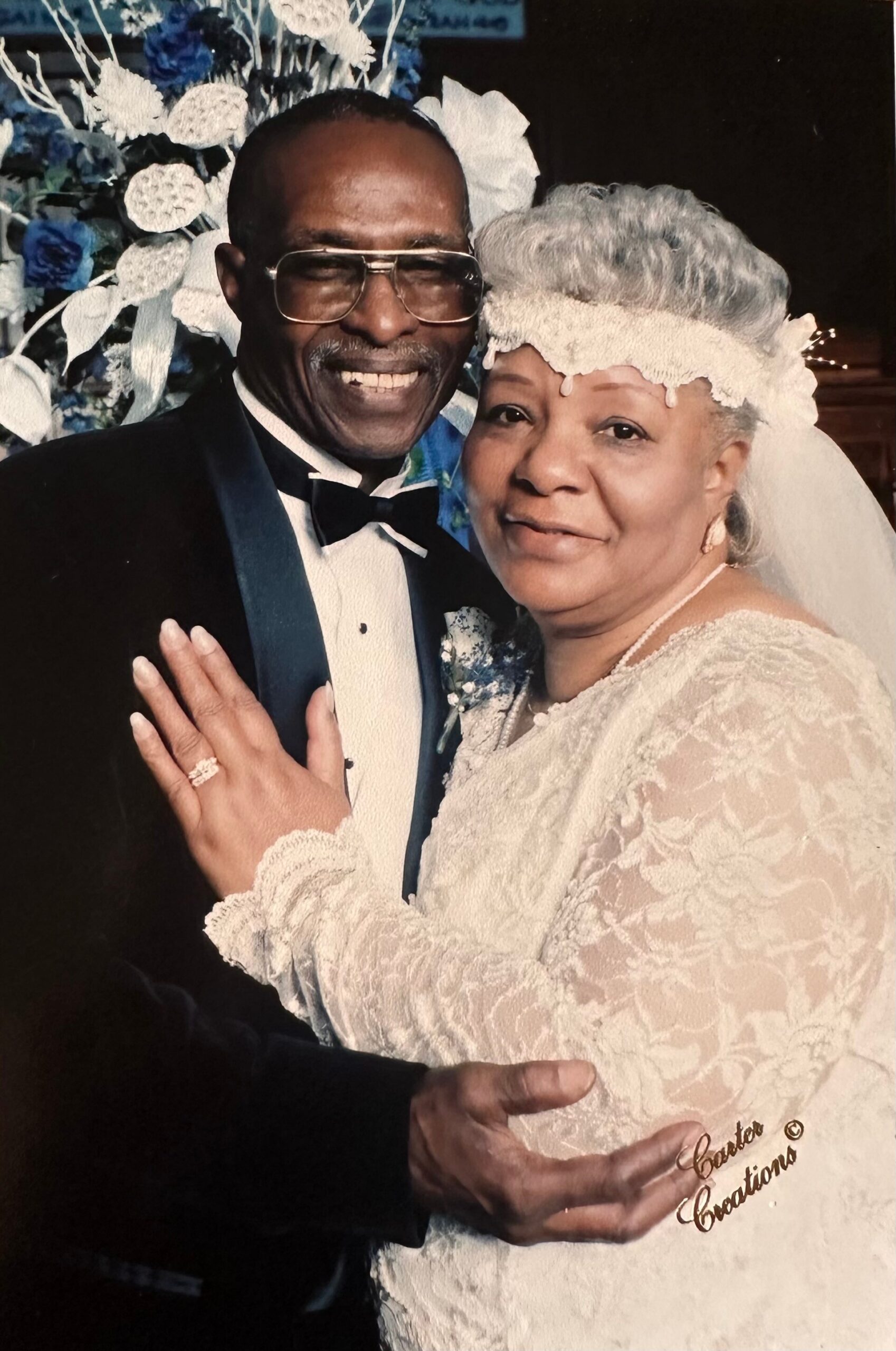 Smiling couple at a wedding: man in a tuxedo with glasses and bow tie, woman in a lace dress with veil and floral headpiece, posing in front of blue-white floral backdrop.