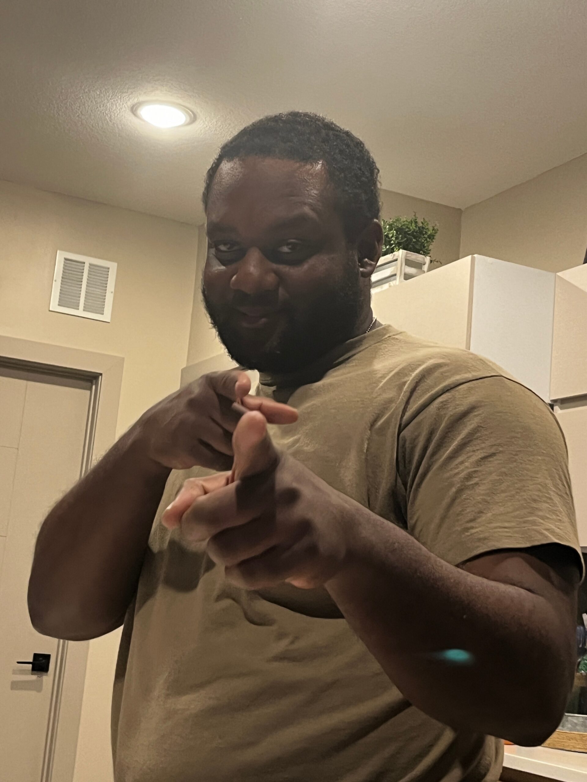 Man in a tan shirt smiling and extending his fists toward the camera in a kitchen setting.