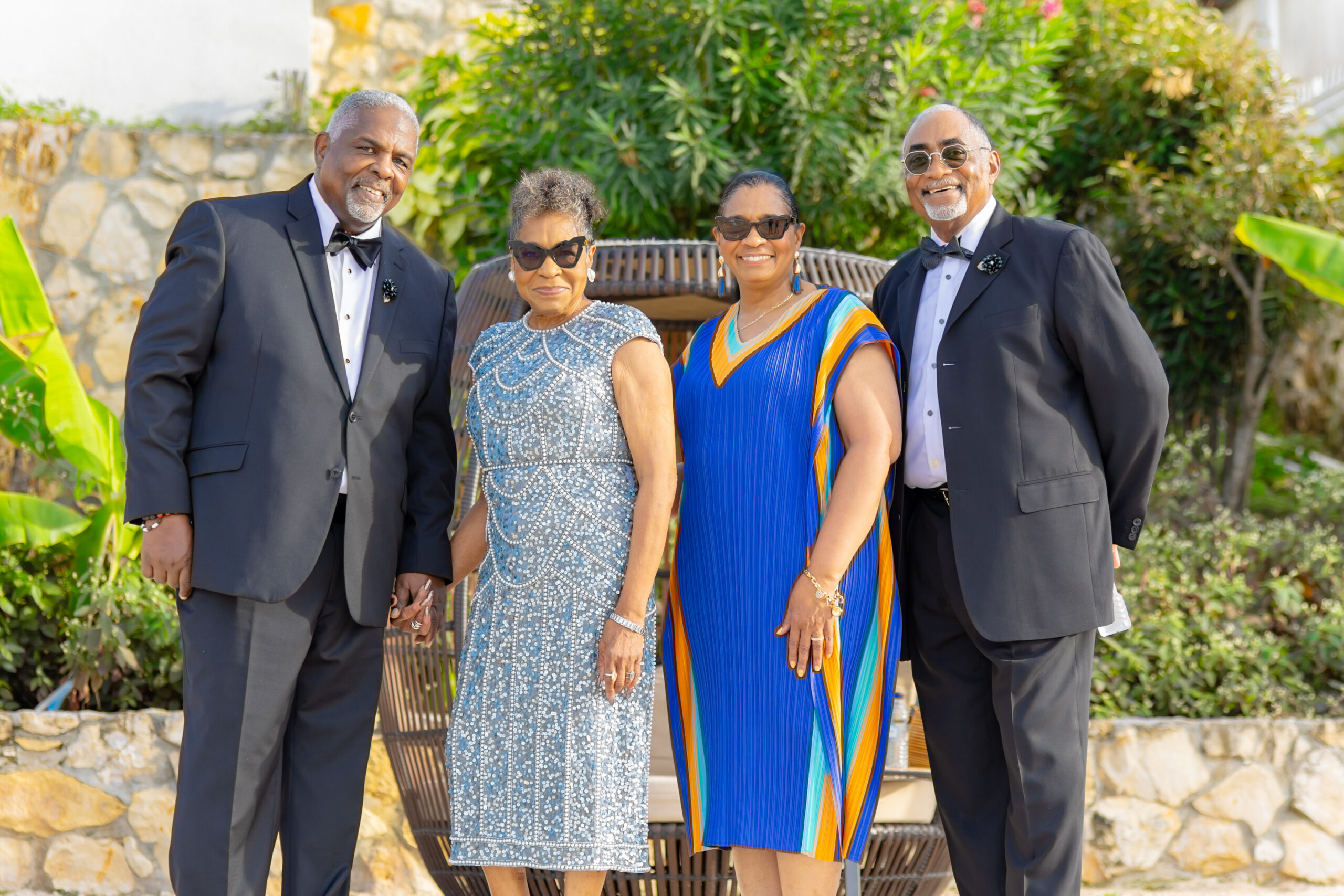 Four adults in formal attire posing outdoors, smiling, with green plants and stone wall in the background.