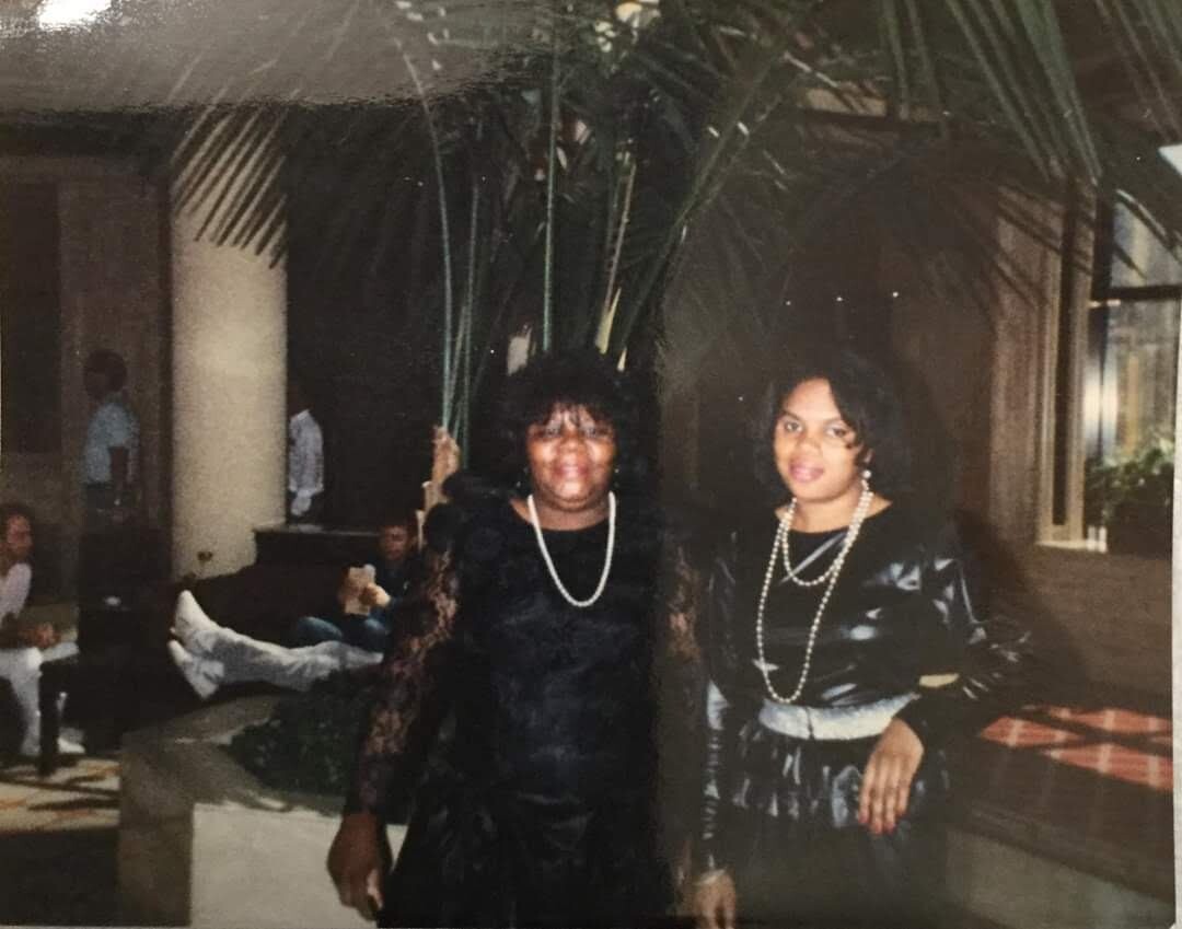 Two women in black dresses with pearl necklaces pose together at a party, with plants and guests in the background.