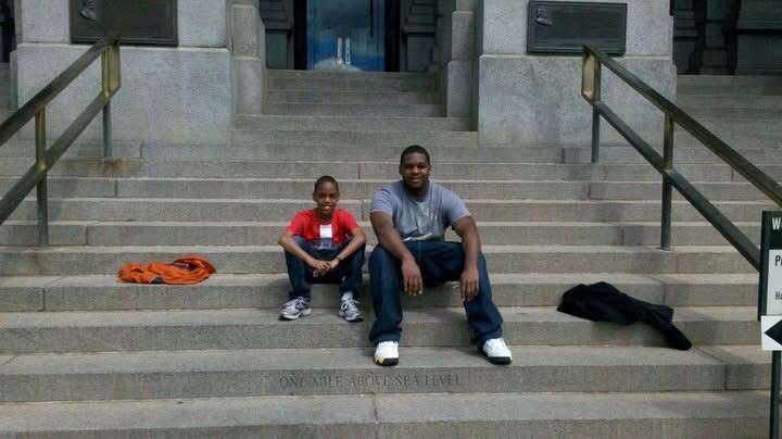 Two men sit on wide stone steps outside a large building; an orange hoodie lies to the left and a black jacket to the right.